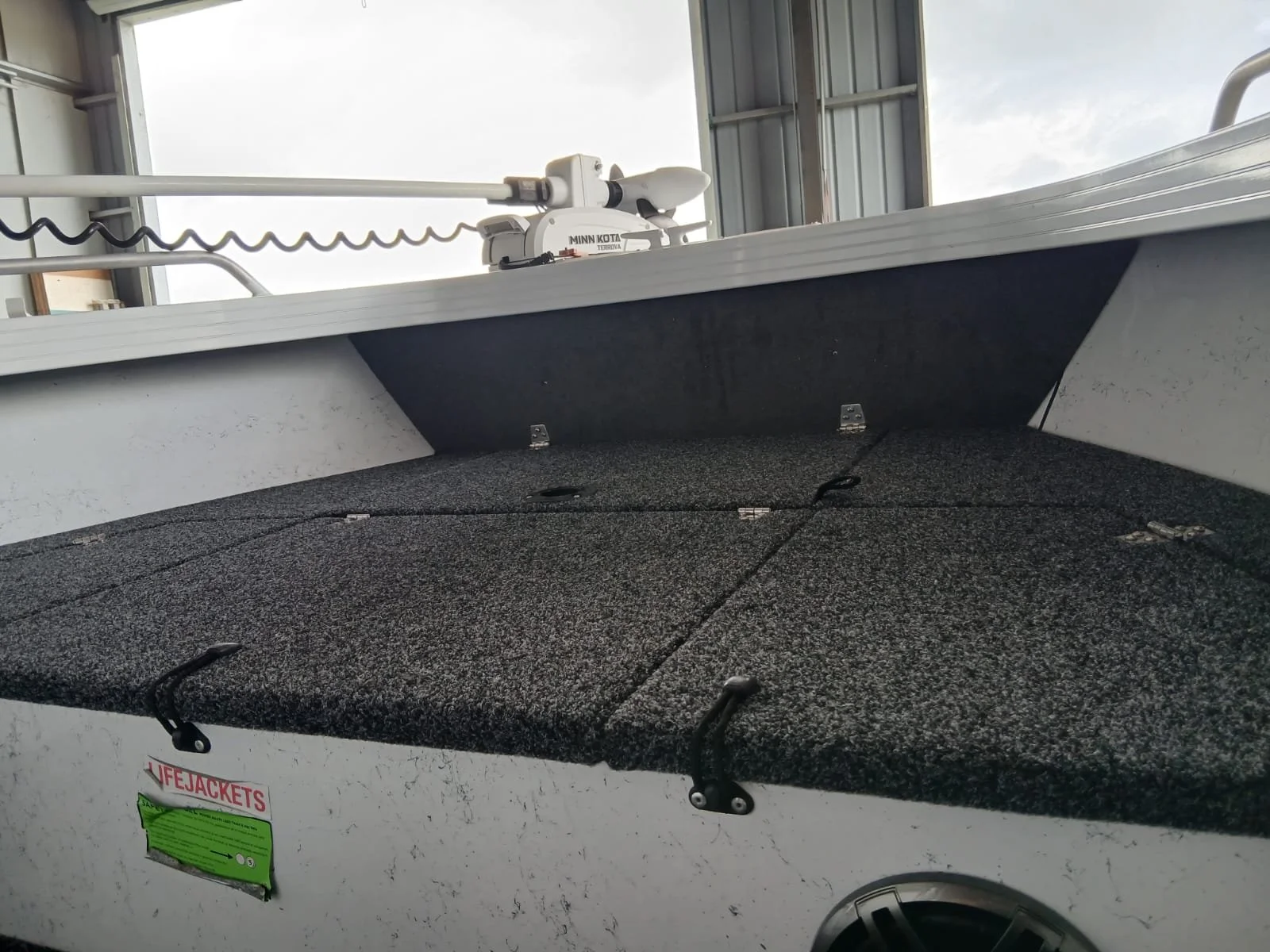 The image shows the inside of a boat with a padded carpeted floor, an open storage compartment, and an electrical motor mounted on the bow with a white pole extending outward.