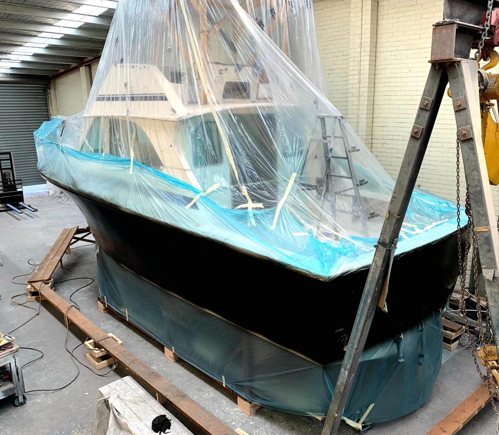 A boat is being covered with a plastic tarp in a workshop, with scaffolding and tools around it.