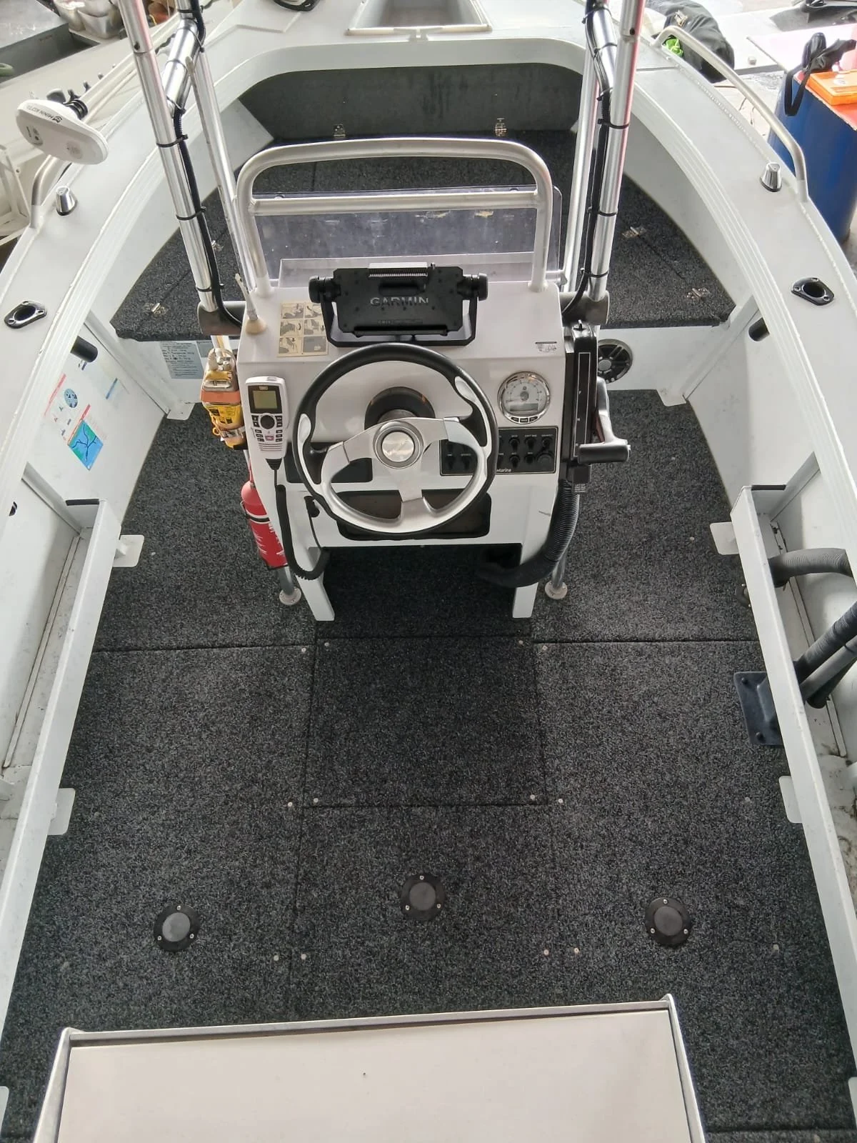 View of the interior of a small boat showing the steering console with a steering wheel, digital display, gauges, and controls. The boat has a black carpeted floor and white sides.