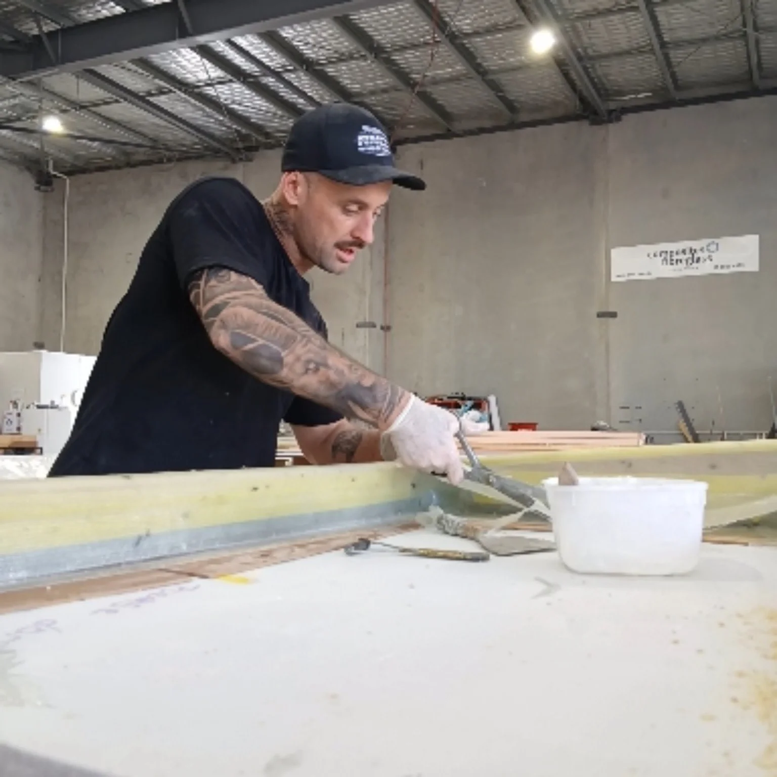 A man with tattoos and a black cap working on a large piece of material in an industrial workshop, using a tool and wearing gloves.