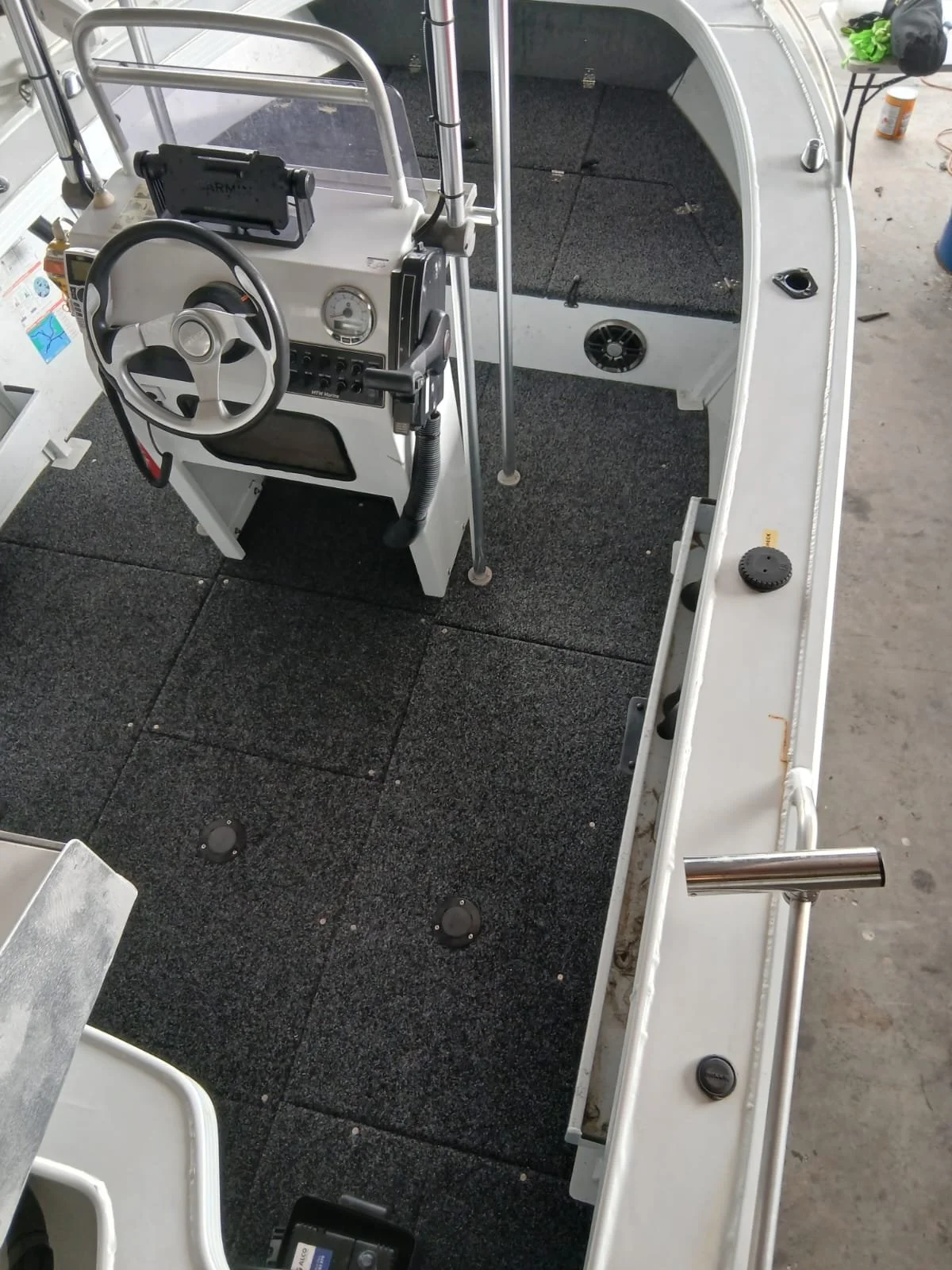 The interior of a small boat showing a white dashboard with a steering wheel, a gauge, various switches, and a black mounted device. The boat has black carpeting on the floor, metal support poles, and a white exterior railing. In the background, we s
