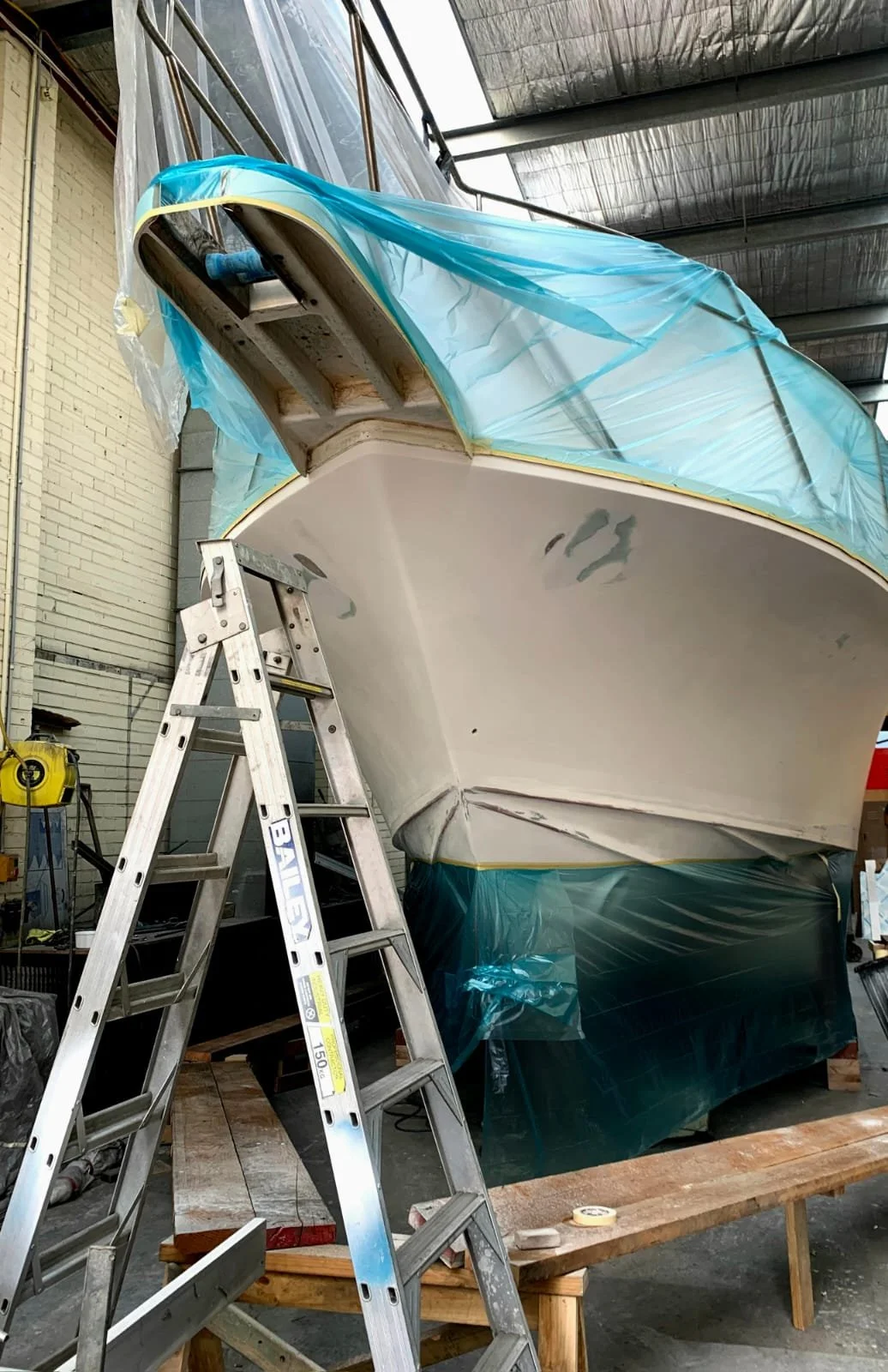 The underside of a boat in a workshop with a ladder leaning against it. The boat is partially covered with blue plastic sheeting.