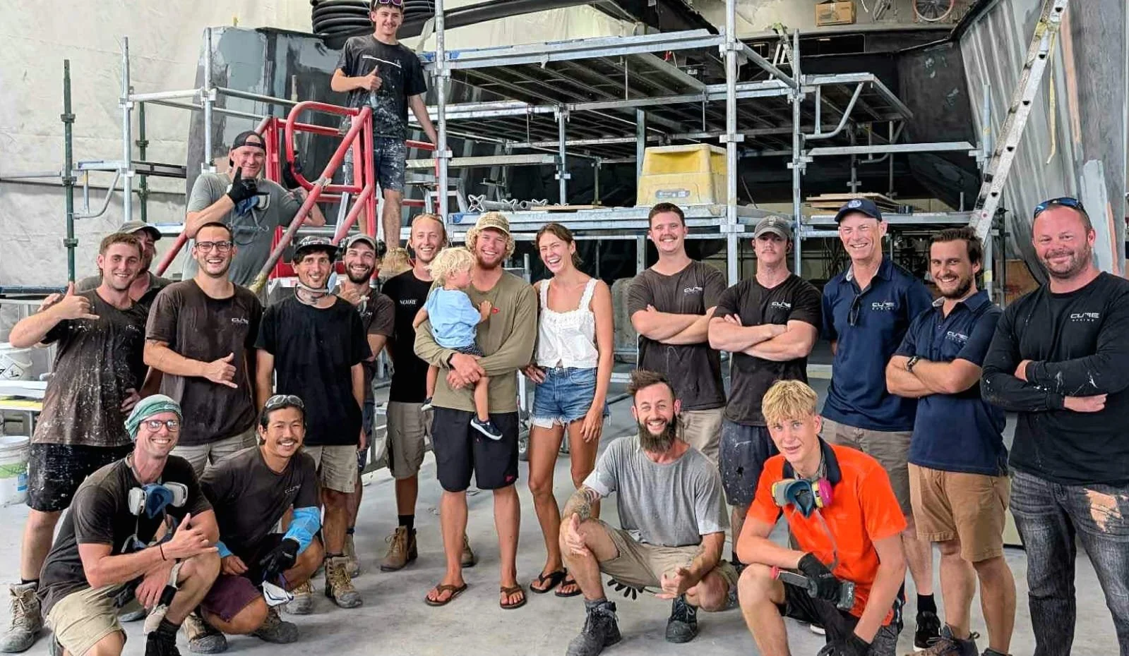 Group of construction workers and a woman with a child, smiling and posing for a photo on a construction site with scaffolding in the background.