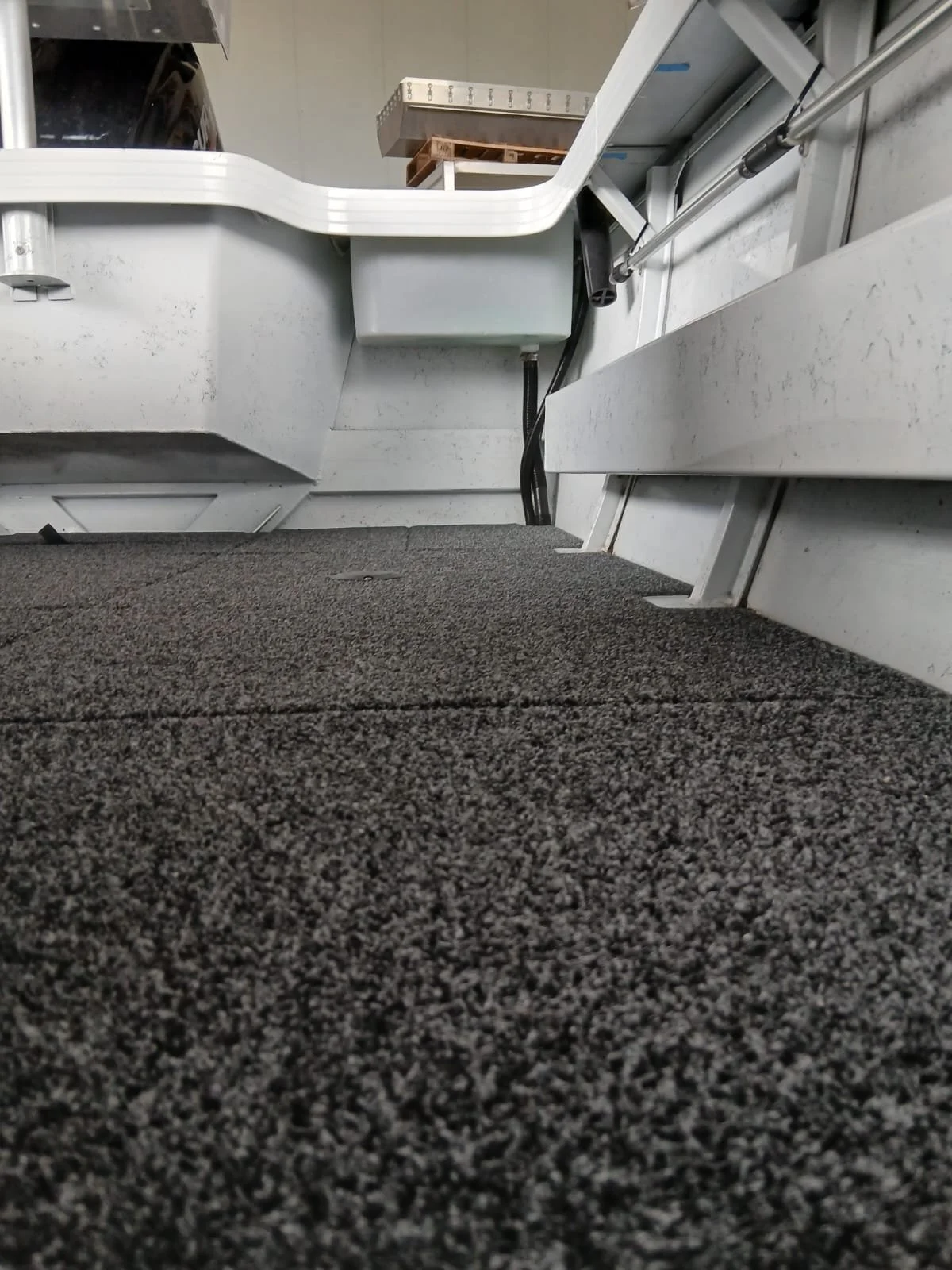 Close-up view of a carpeted floor with industrial furniture and equipment, including pipes and a grey box, inside a warehouse or industrial setting.