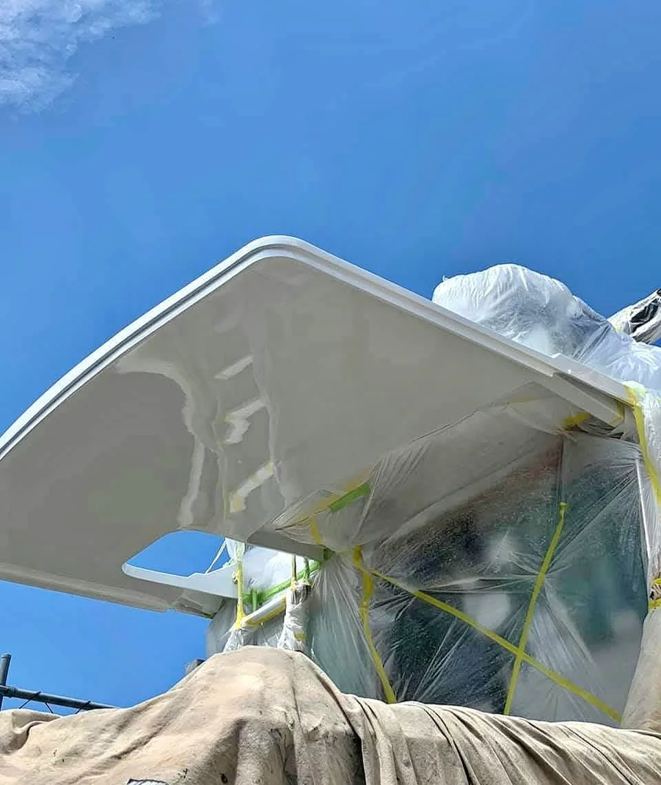 Close-up view of a construction or renovation project showing a section of a white ceiling being painted or coated, with protective plastic sheeting and yellow tape, against a clear blue sky.