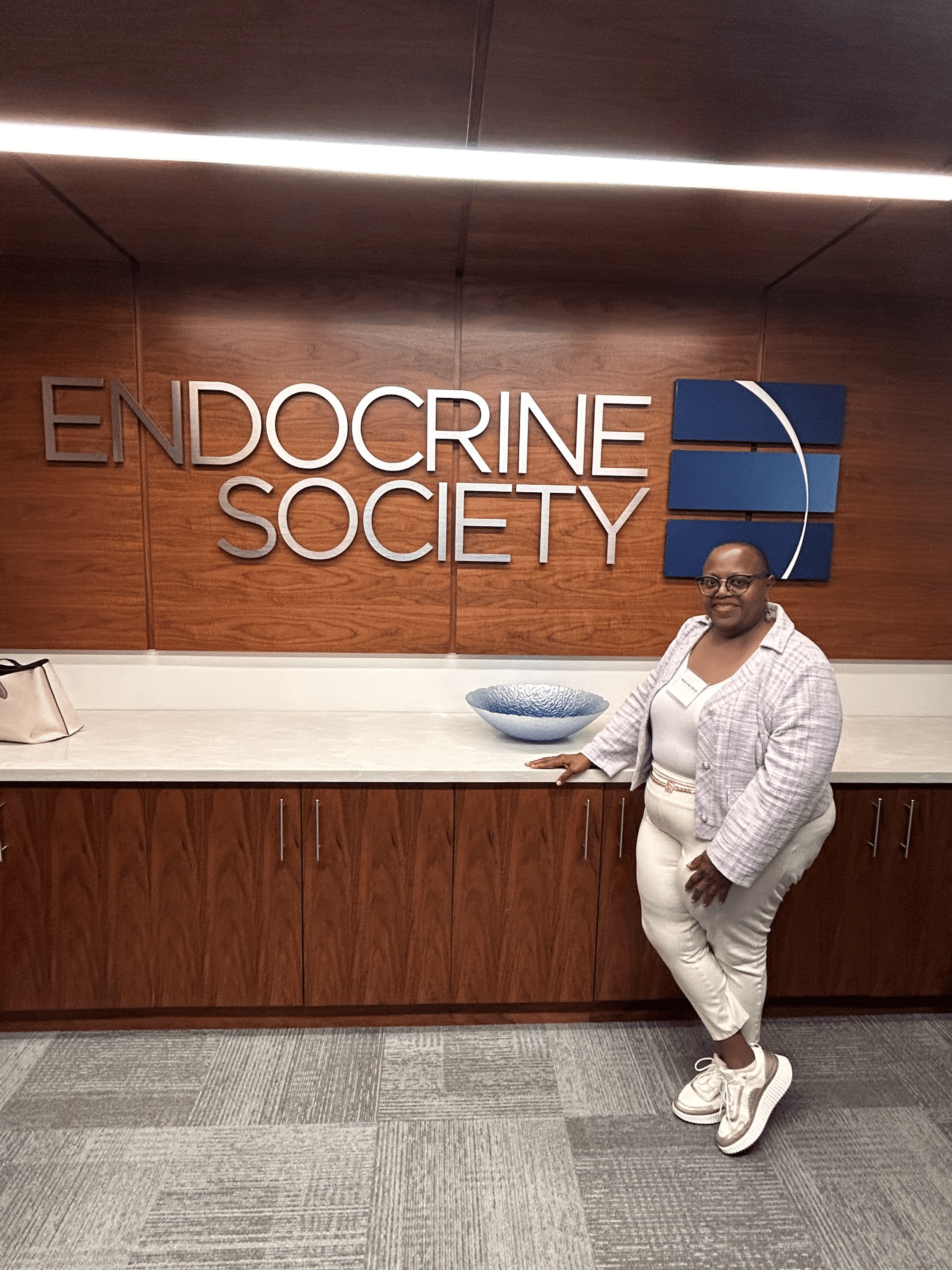 A woman smiling and leaning against a wooden counter in front of a wooden wall with a sign that reads 'Endocrine Society' and a blue logo. She is wearing white pants, a light plaid blazer, and white sneakers.