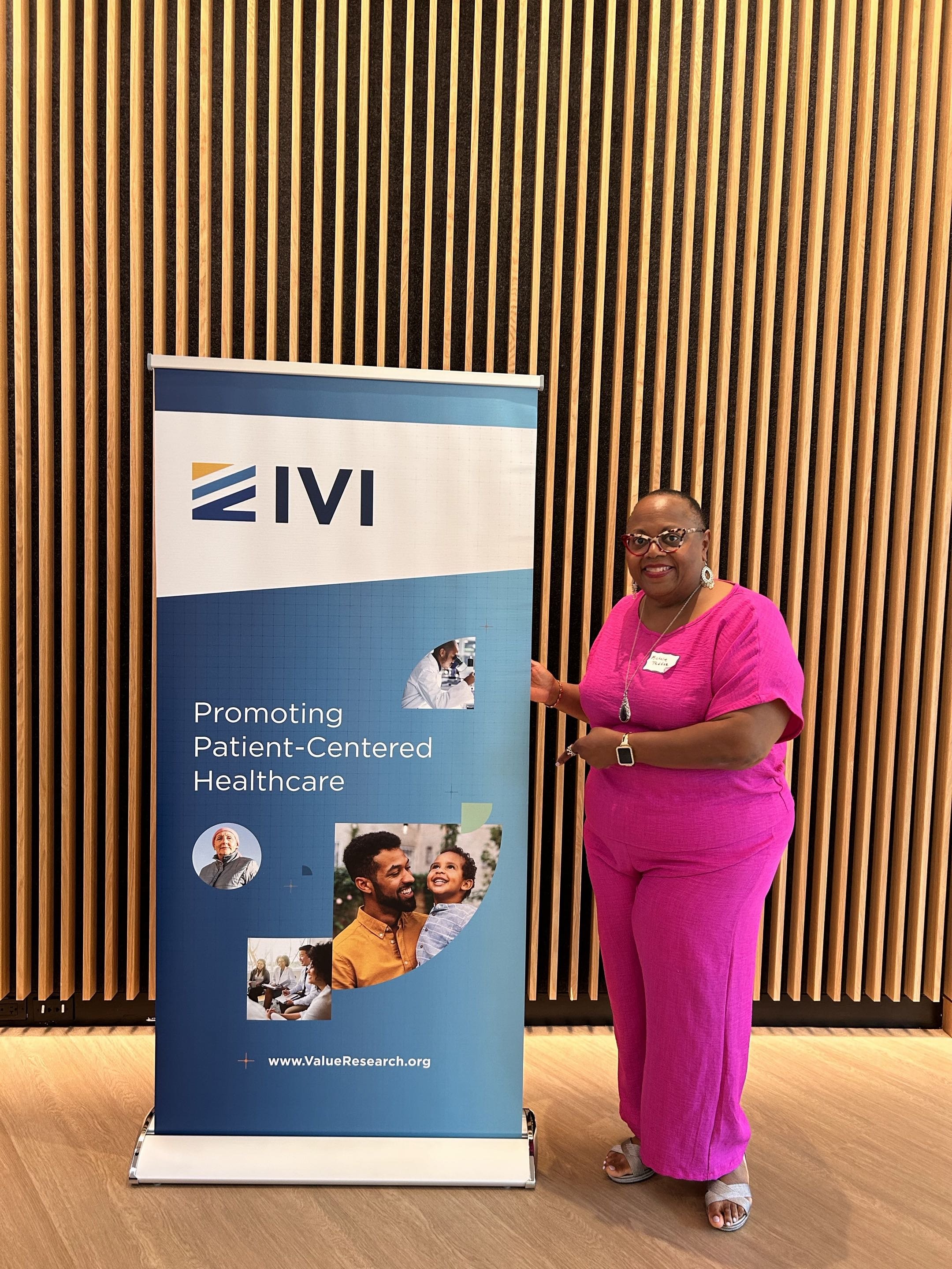 A woman in a pink outfit standing next to a vertical banner with the logo VI and the text "Promoting Patient-Centered Healthcare". She is smiling and wearing glasses, earrings, a watch, and a necklace, with a wooden slat wall in the background.