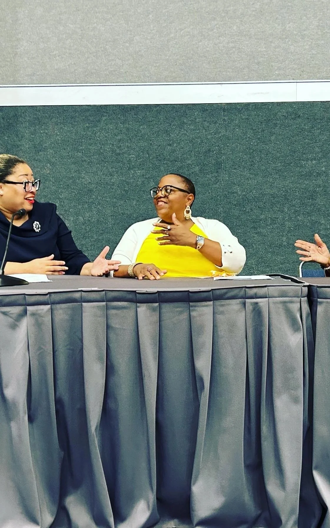 Three women sitting at a table engaged in conversation, with a green wall in the background and gray curtain in front.