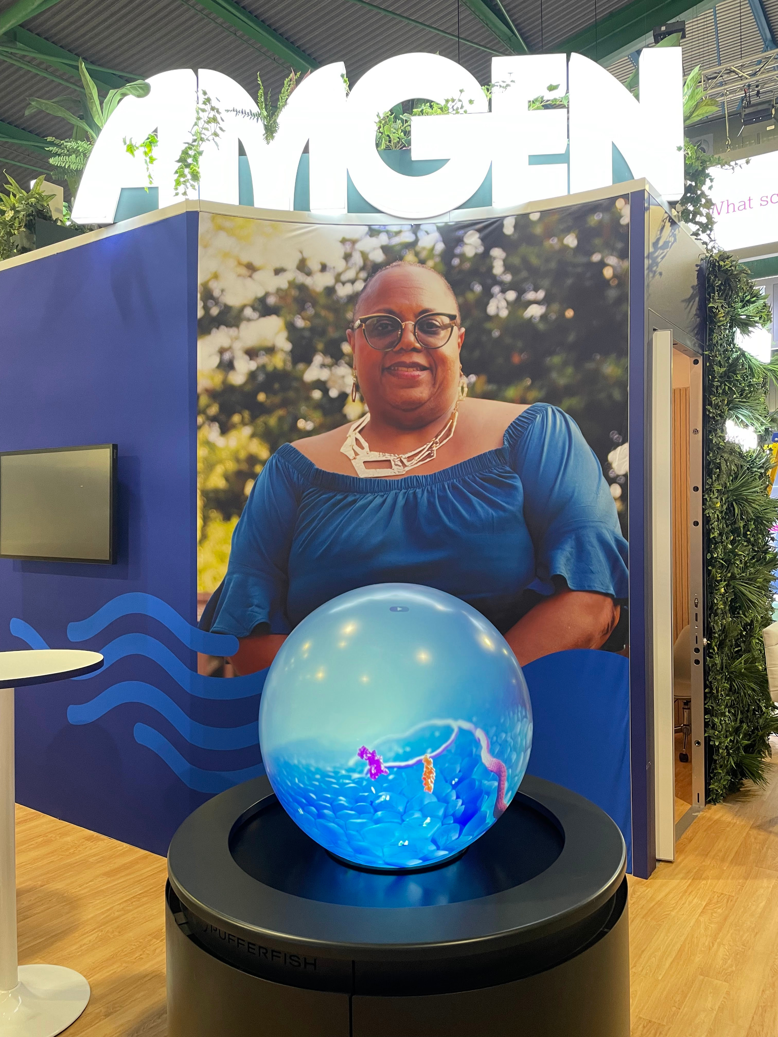 A person with glasses and earrings wearing a blue top, smiling, standing in front of an aquarium display with a blue background and a large fish illustration. The display is part of an exhibition or event space with a sign that says 'AMGEN' above, and green plants surrounding it.