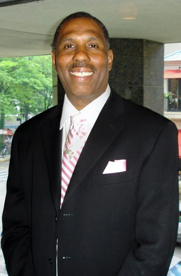 Smiling man in a black suit with a white shirt, pink striped tie, and pink pocket square, standing indoors with a window showing green trees in the background.