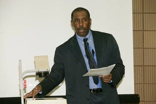 Man in a dark suit giving a presentation, holding papers, standing in front of a whiteboard.