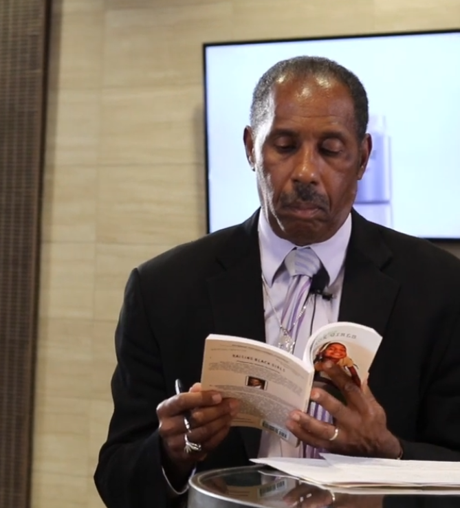 A man in a suit and tie reading a booklet at a table in a room with a beige wall and a large screen in the background.