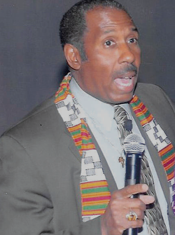 Man speaking into a microphone, wearing a gray suit and a colorful kente cloth sash.
