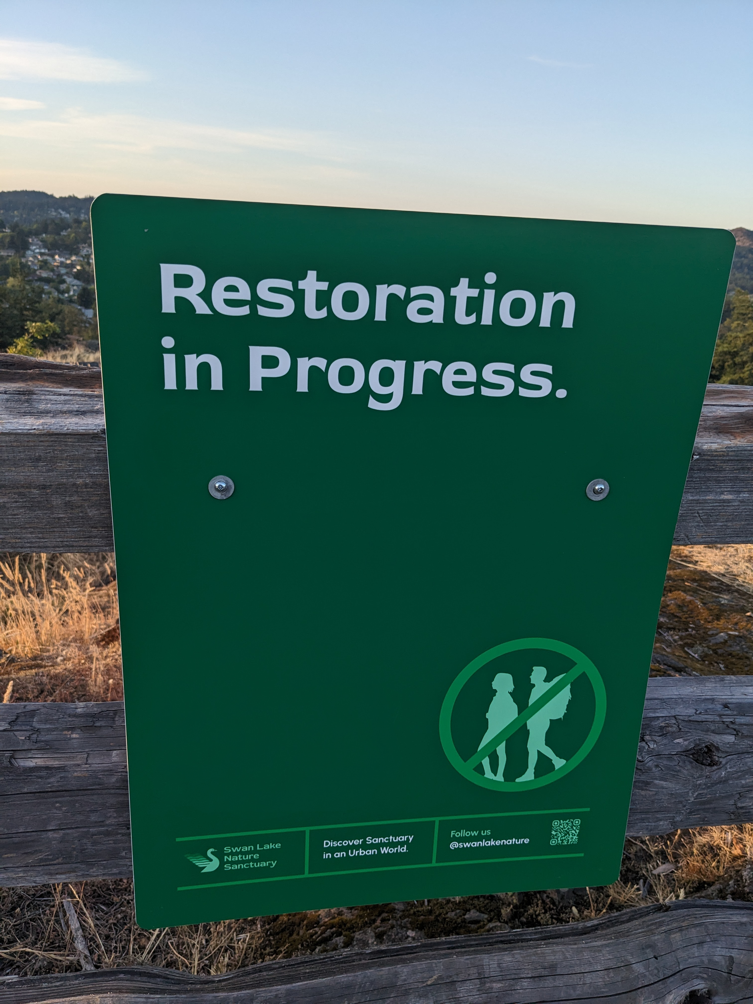 A green sign saying 'restoration in progress' attached to a fence overlooking a large nature area.
