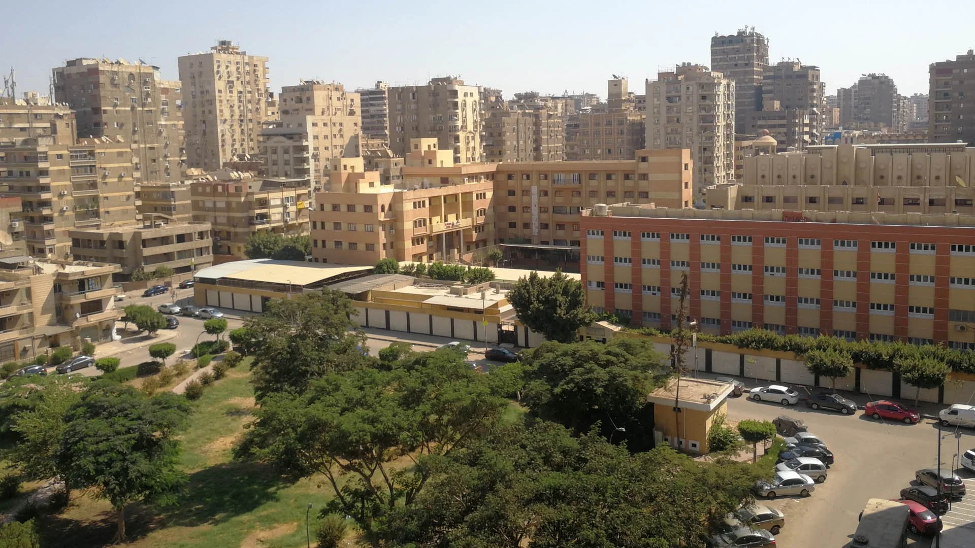 Cairo cityscape with tall beige apartment buildings, some with colorful accents, green trees and a parking lot with cars in the foreground.