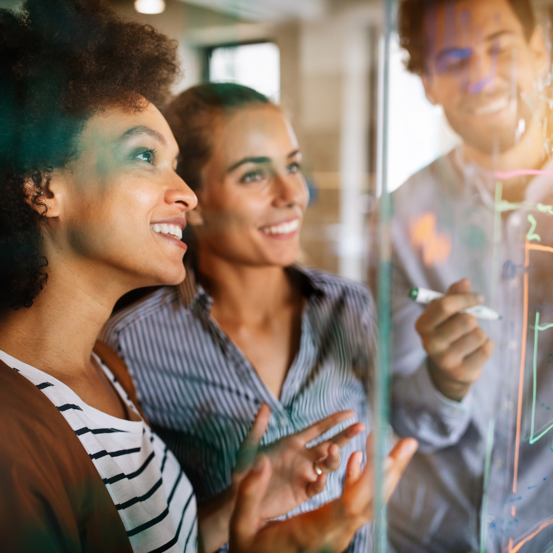 Three employees collaborating on a project, smiling and discussing while writing on a transparent board with colorful diagrams.