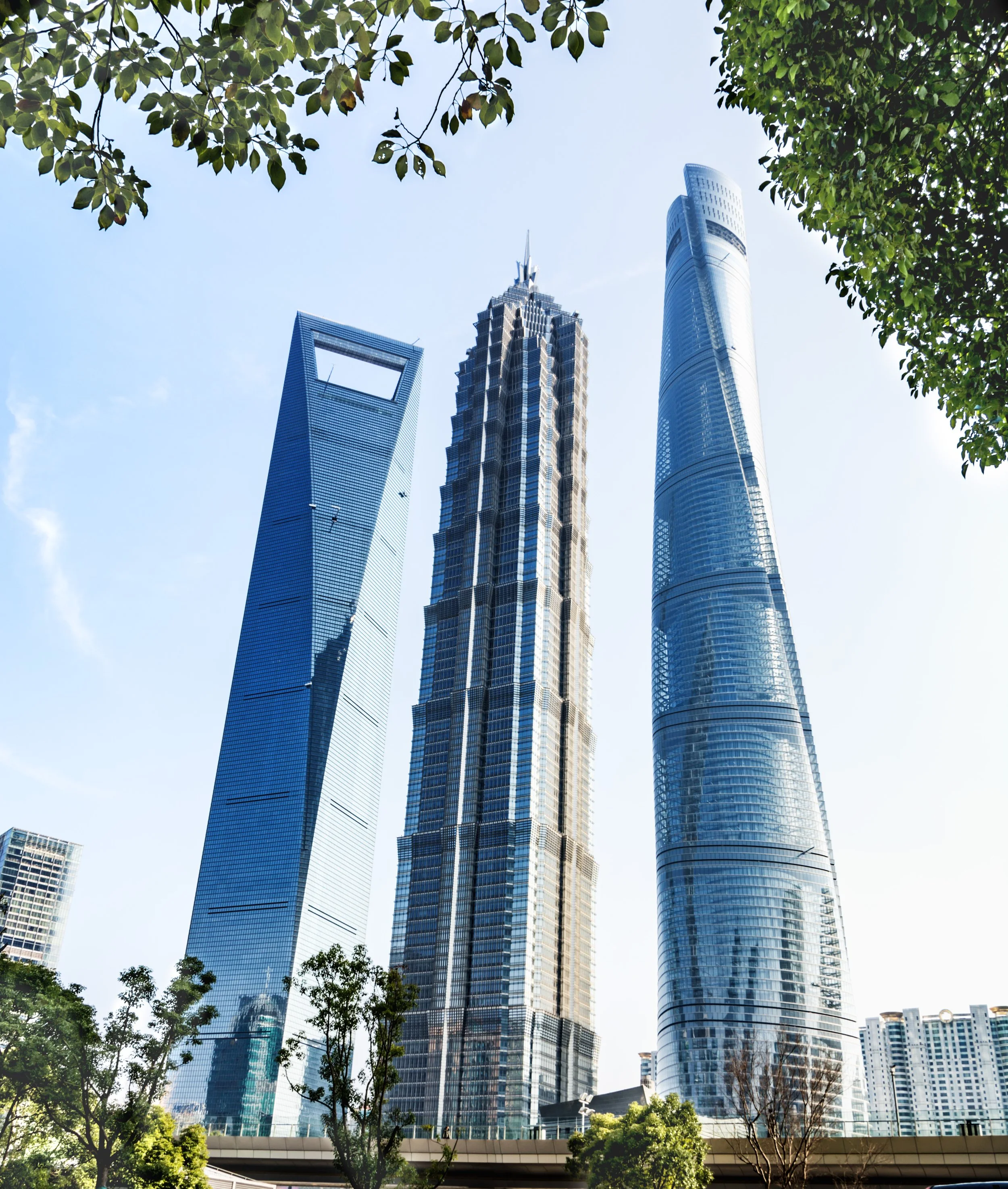 Riyadh skyline with three modern skyscrapers with glass facades, framed by leafy trees in the foreground, under a clear blue sky.