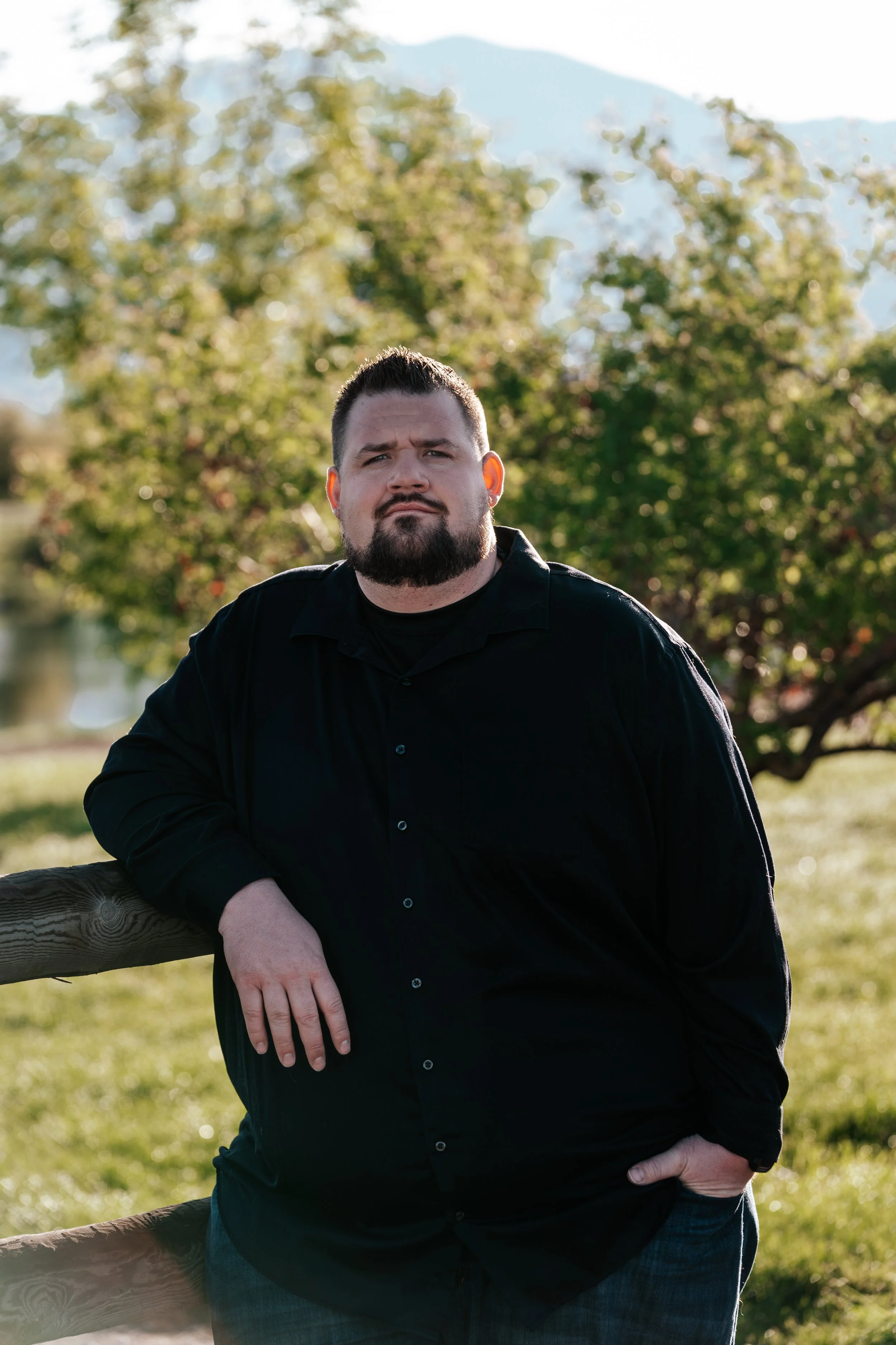 A man with a beard and short dark hair, wearing a black button-up shirt, leaning on a wooden fence outdoors with trees and mountains in the background.