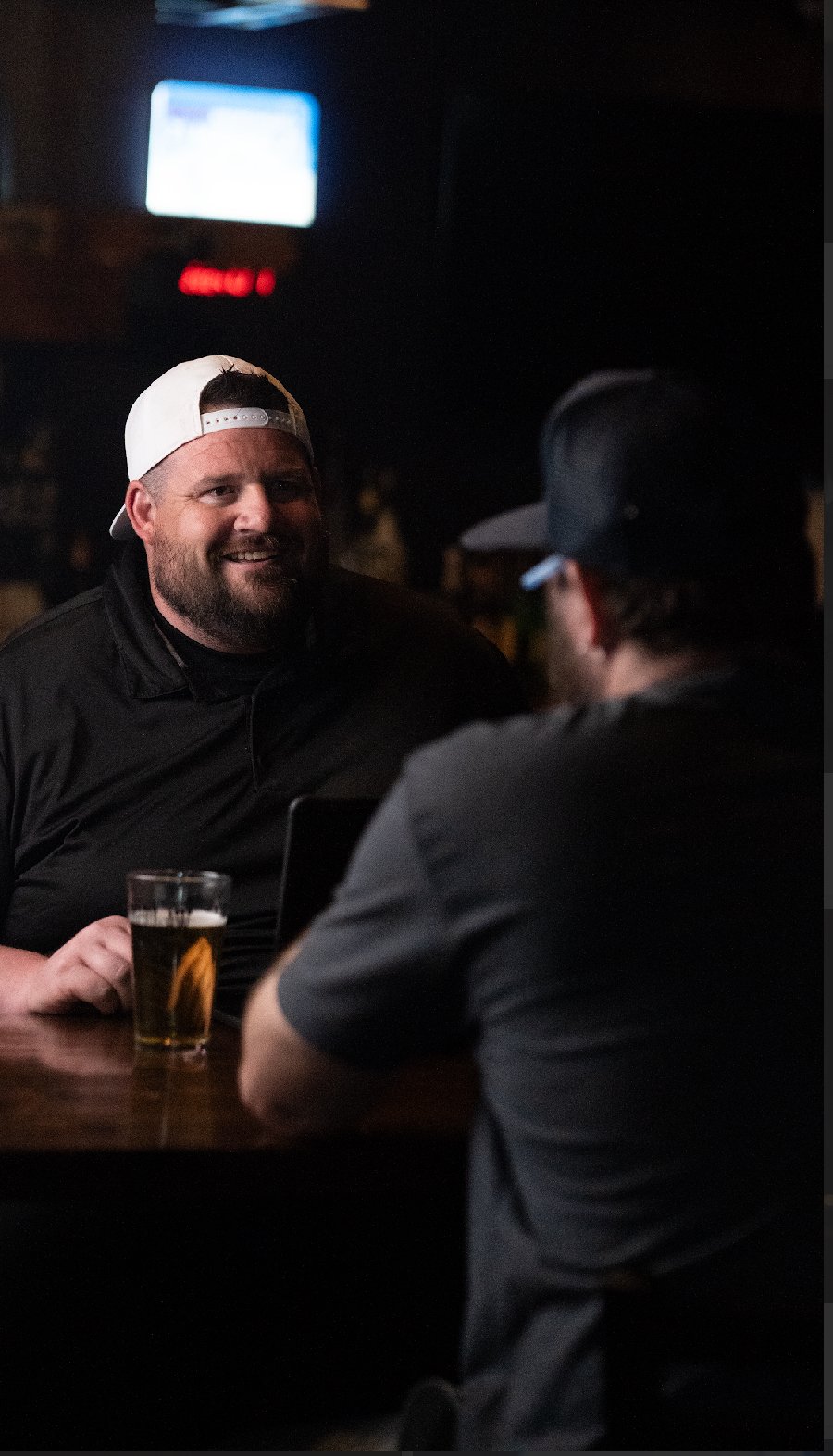 Two men sitting at a bar having a conversation, one smiling, with a pint of beer in front of him, in a dimly lit setting.