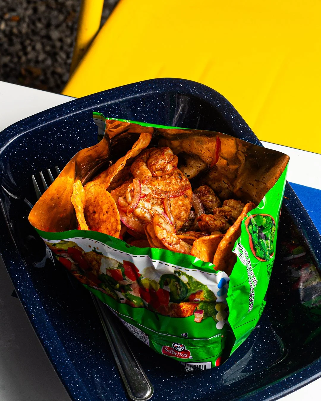 Open bag of mixed chips and seasoned seafood in black tray with yellow background.