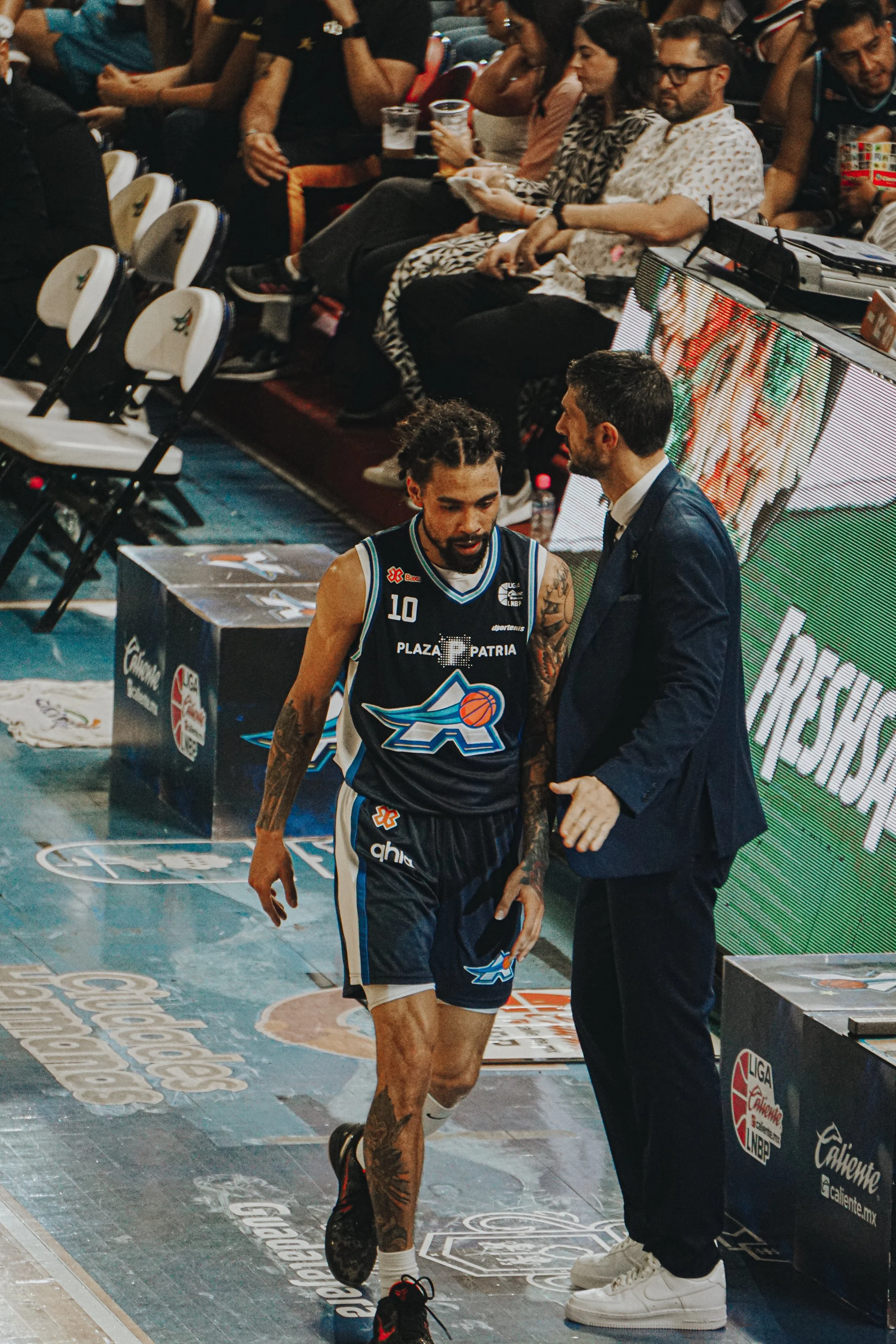 A basketball player with tattoos on his arm and leg, wearing a black jersey with the number 10, talking to his coach on the sideline during a game, with spectators seated behind them.
