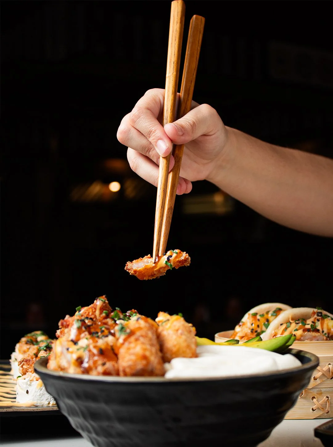 A hand using chopsticks lifting a piece of fried food from a bowl of Asian cuisine. The dish contains fried items garnished with green herbs and accompanied by white sauce. There are also noodles with sauce in the background.