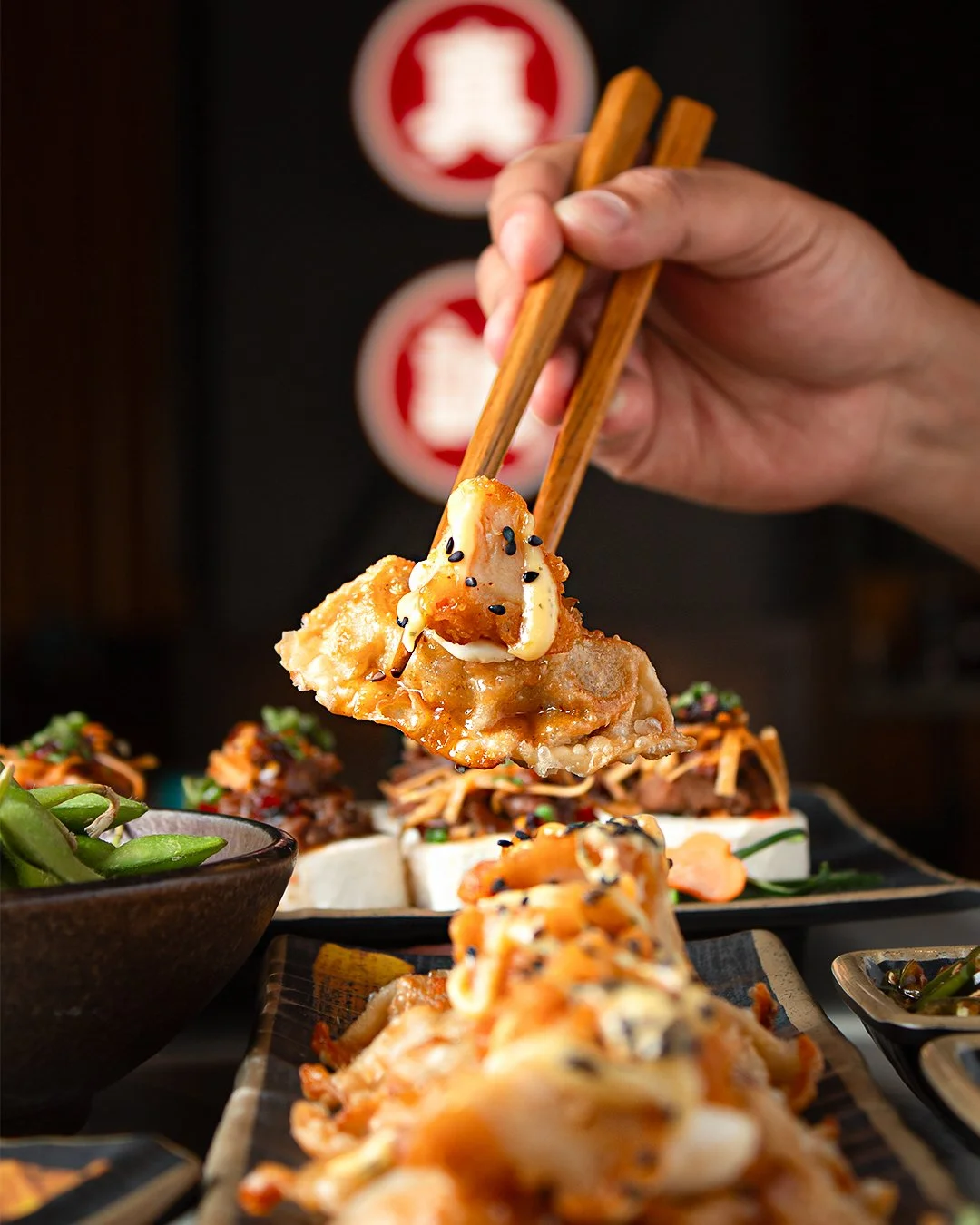 Close-up of a hand holding chopsticks picking up a piece of glazed chicken garnished with black sesame seeds, with various Asian dishes on the table in the background.