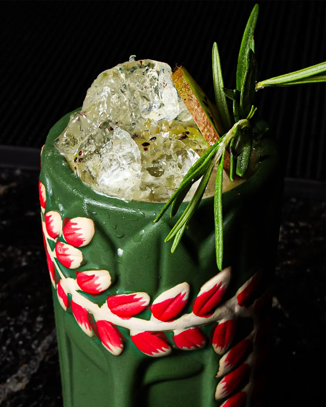 A cocktail in a cactus-shaped mug garnished with rosemary and a slice of apple, filled with ice cubes.