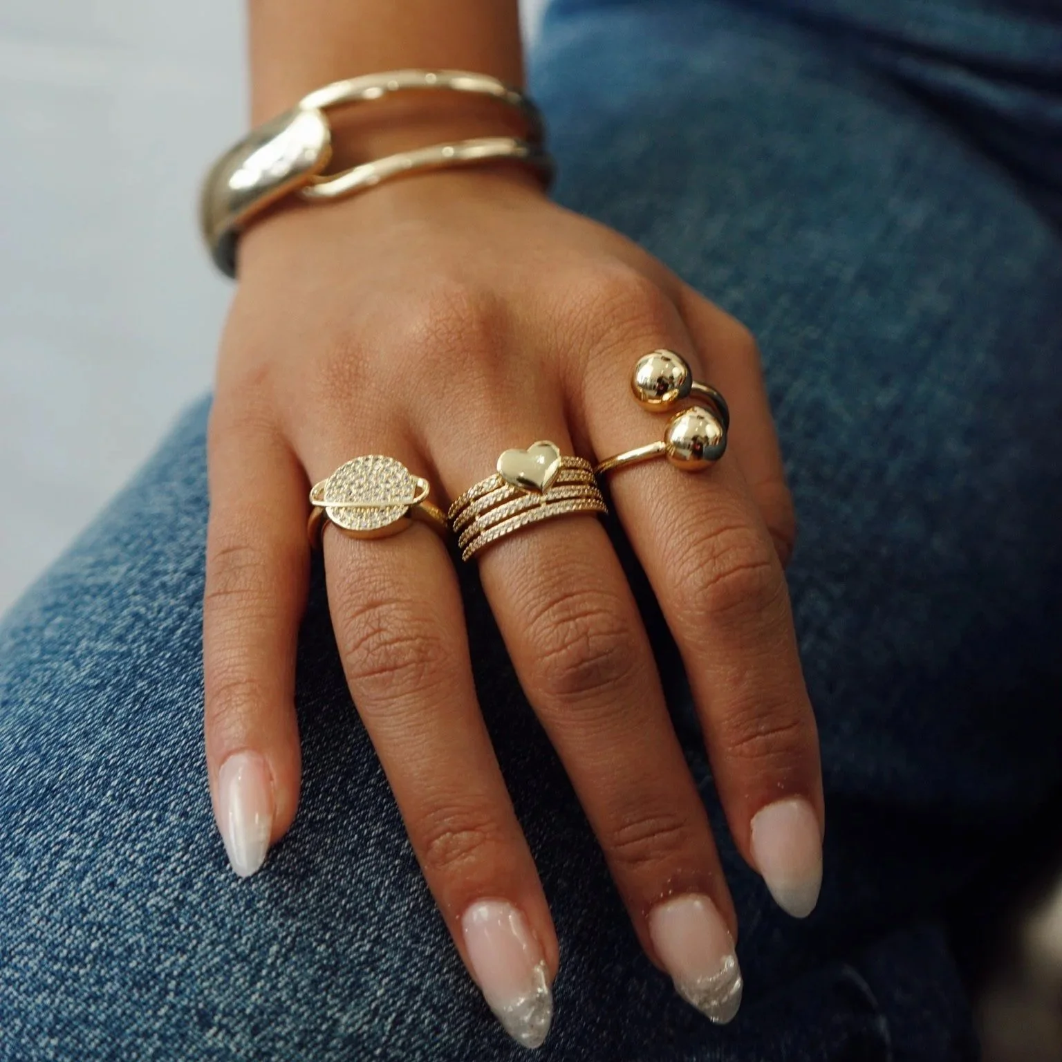 A person's hand resting on their jeans, adorned with multiple gold rings and bracelets.