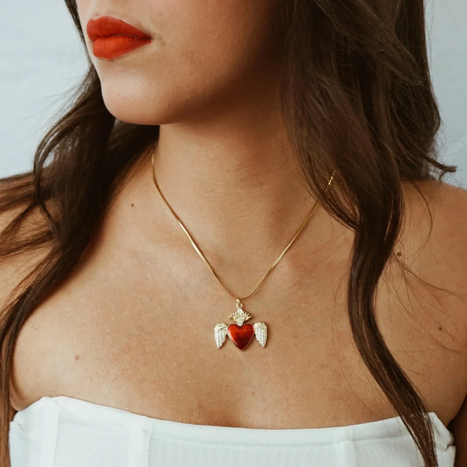 Close-up of a woman wearing red lipstick and a gold necklace with a pendant featuring a red heart with wings.