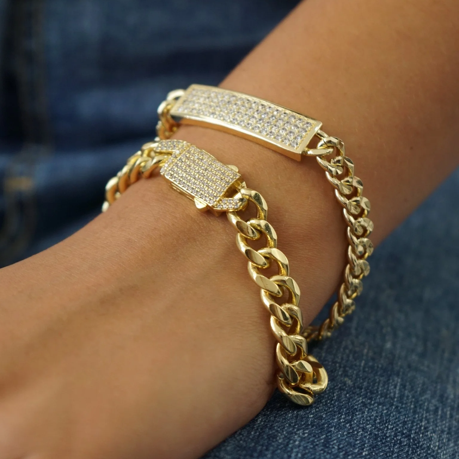 Close-up of a person's wrist wearing two gold chain bracelets with rectangular, rhinestone-studded clasps, against a background of denim fabric.