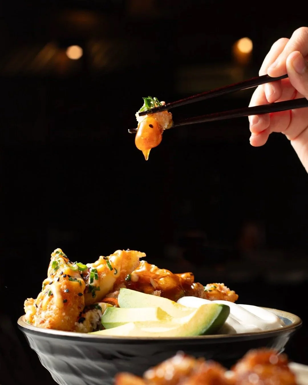 A hand holding chopsticks with a piece of sushi above a bowl of sushi, fried items, lime wedges, and sauce.