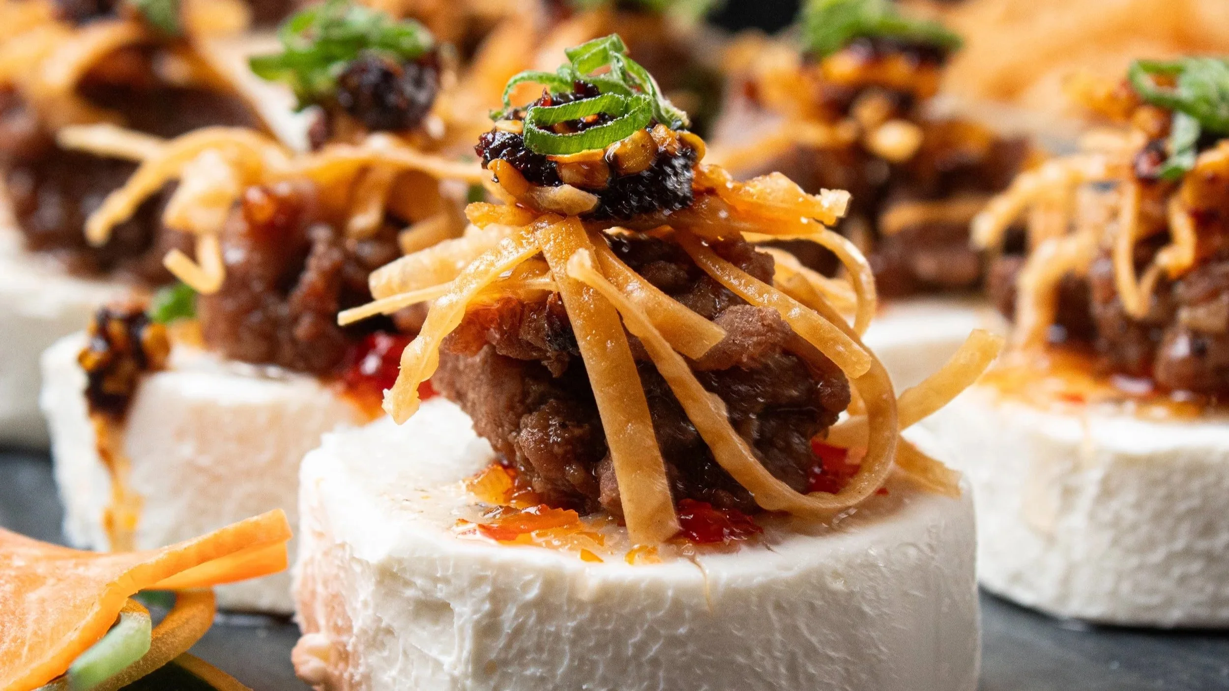 Appetizer of small savory bites featuring a white base topped with slow-cooked beef, shredded cheese, chili, and chopped green onions.