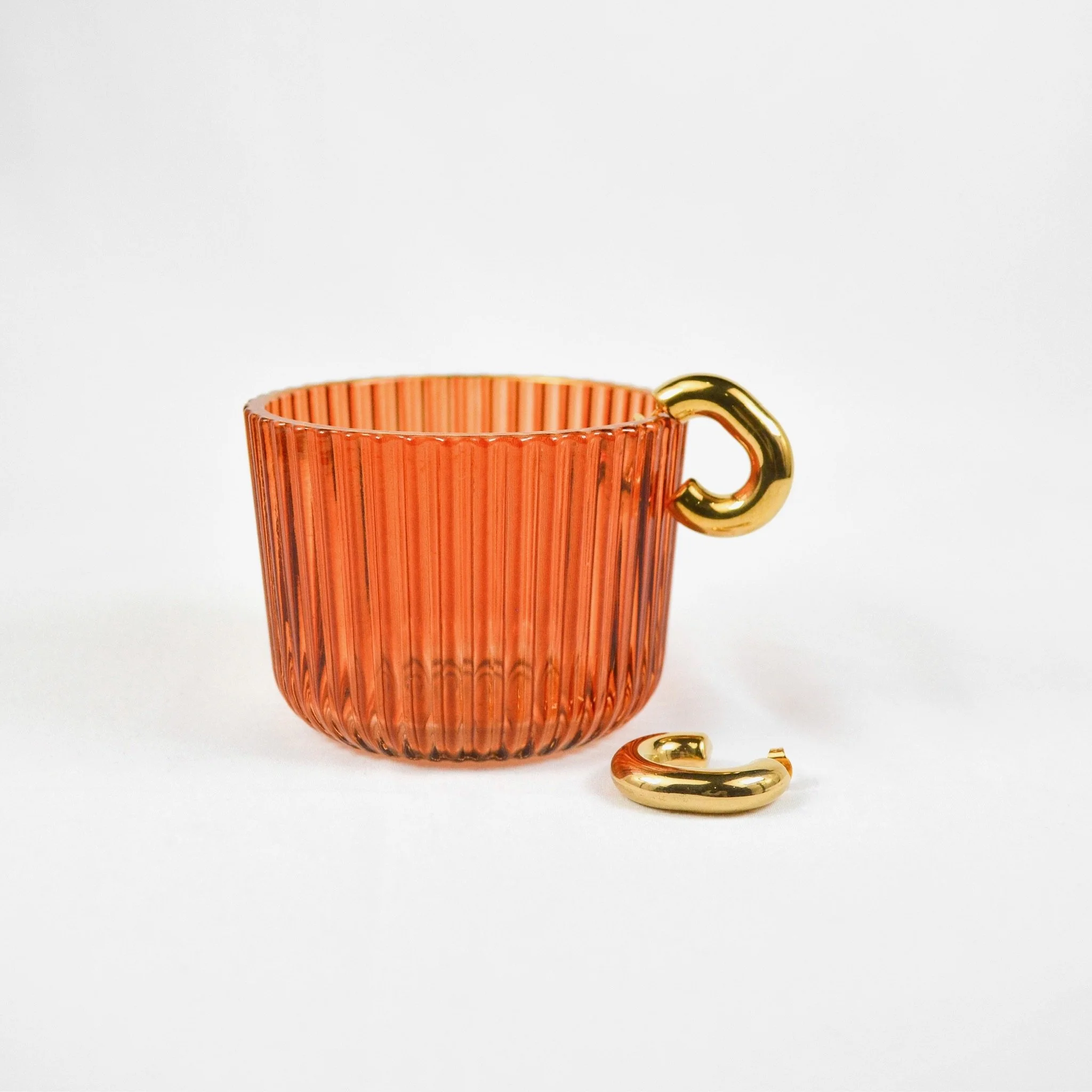 A pink glass cup with vertical ridges and gold-colored heart-shaped handle, with a separate gold-colored heart-shaped handle piece beside it, on a plain white background.