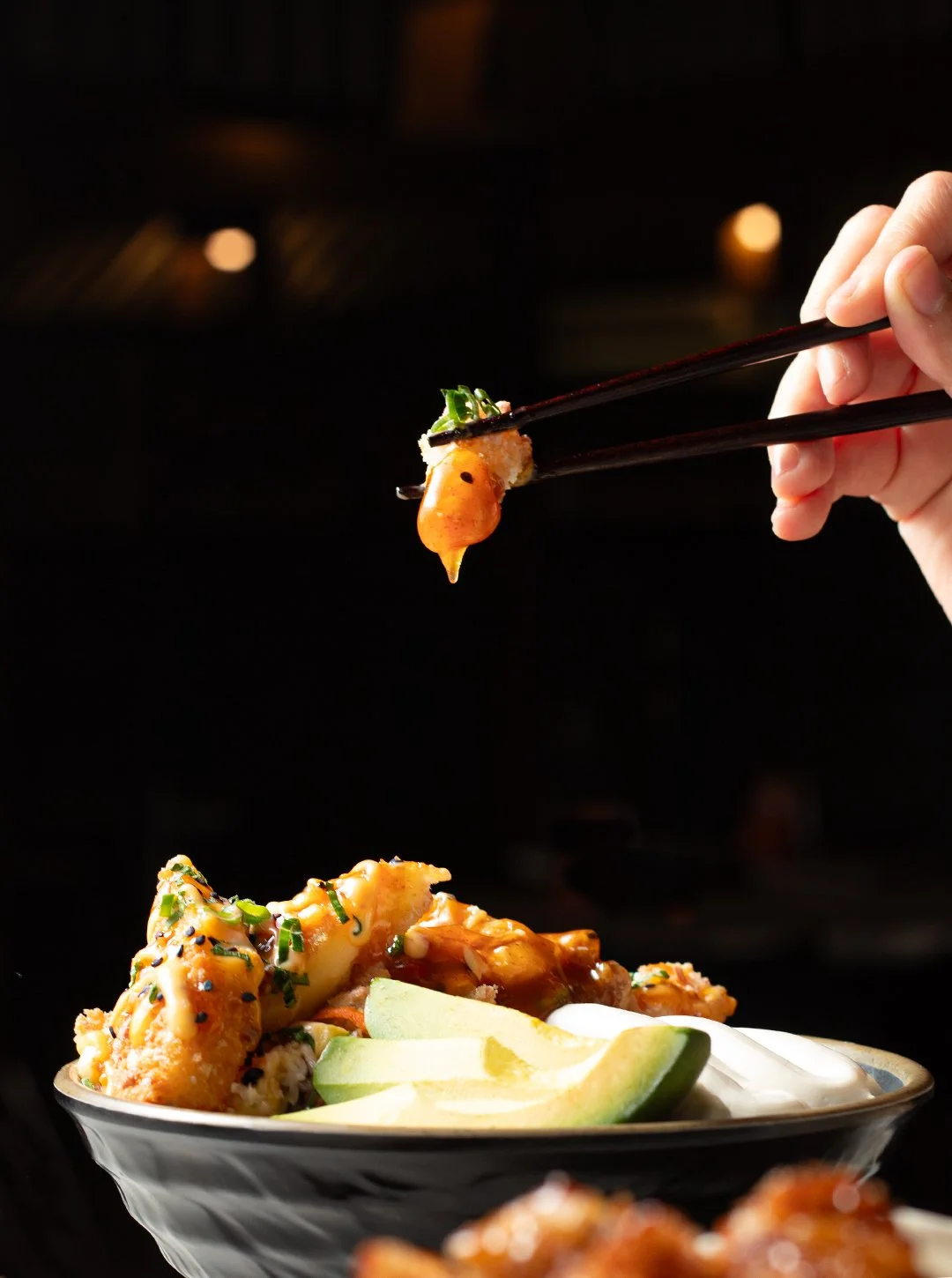 A hand holding chopsticks with a piece of sushi above a bowl of sushi, fried items, lime wedges, and sauce.