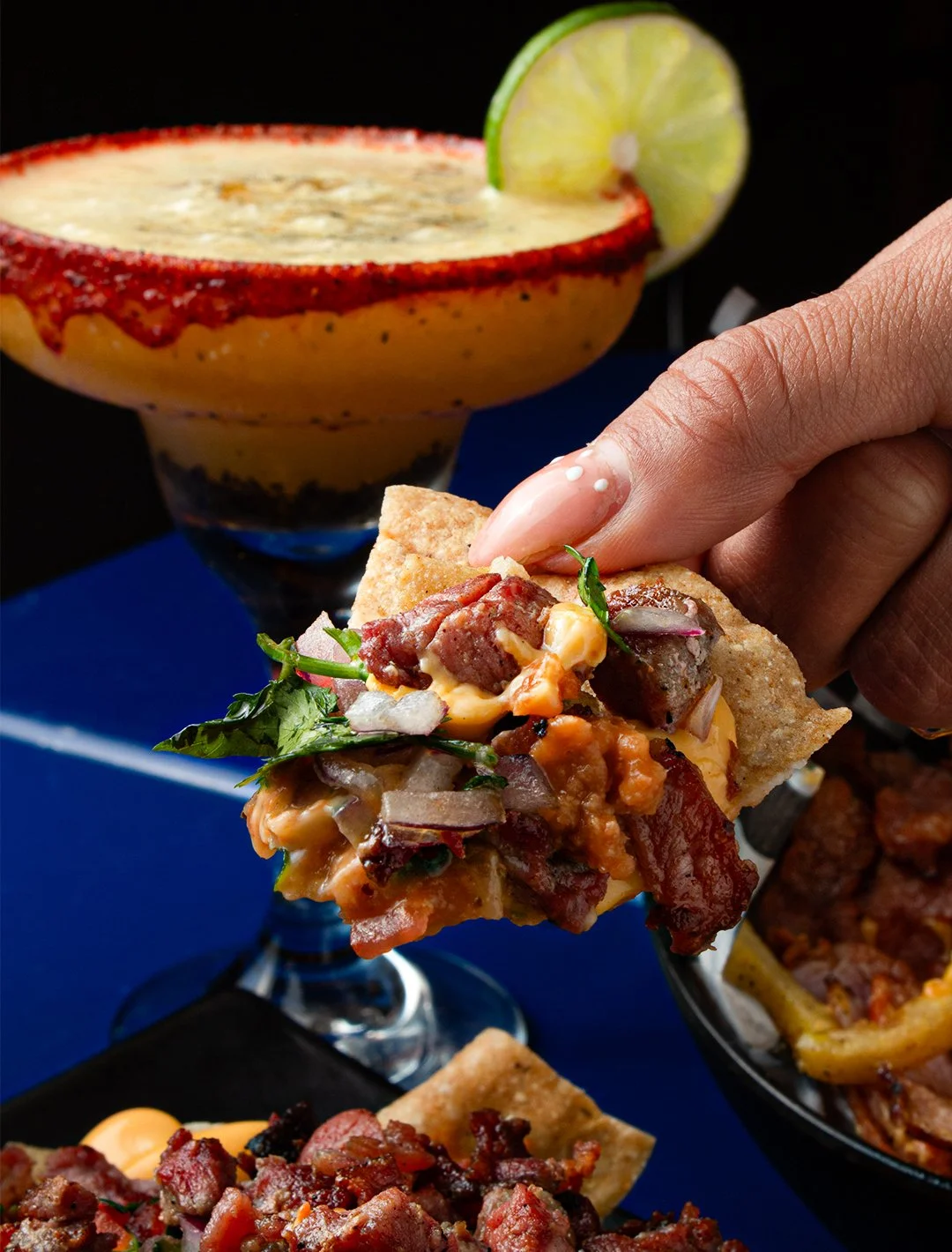 A hand holding a taco with meat, beans, and vegetables, with a lime wedge in the background and a Mexican cocktail in a glass with sugar rim and lime.