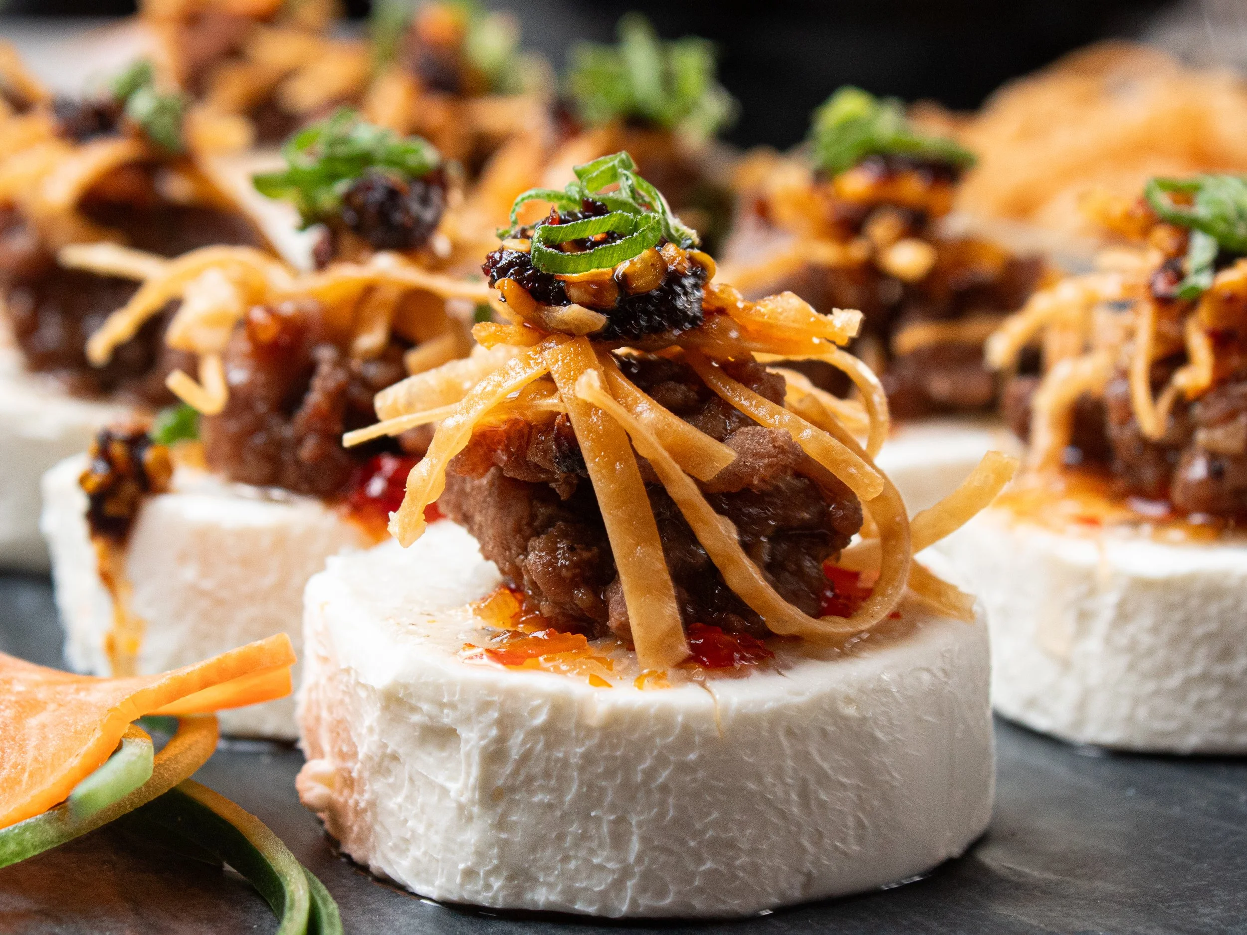 Appetizer of small savory bites featuring a white base topped with slow-cooked beef, shredded cheese, chili, and chopped green onions.