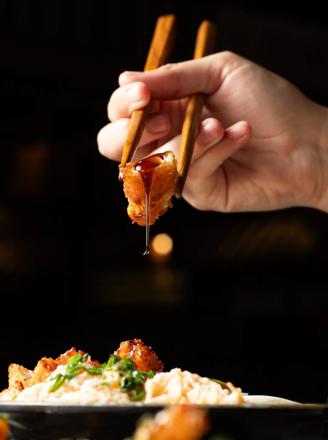 A hand holding chopsticks is dipping a piece of fried food with sauce over a plate of food, including mashed potatoes and green herbs.