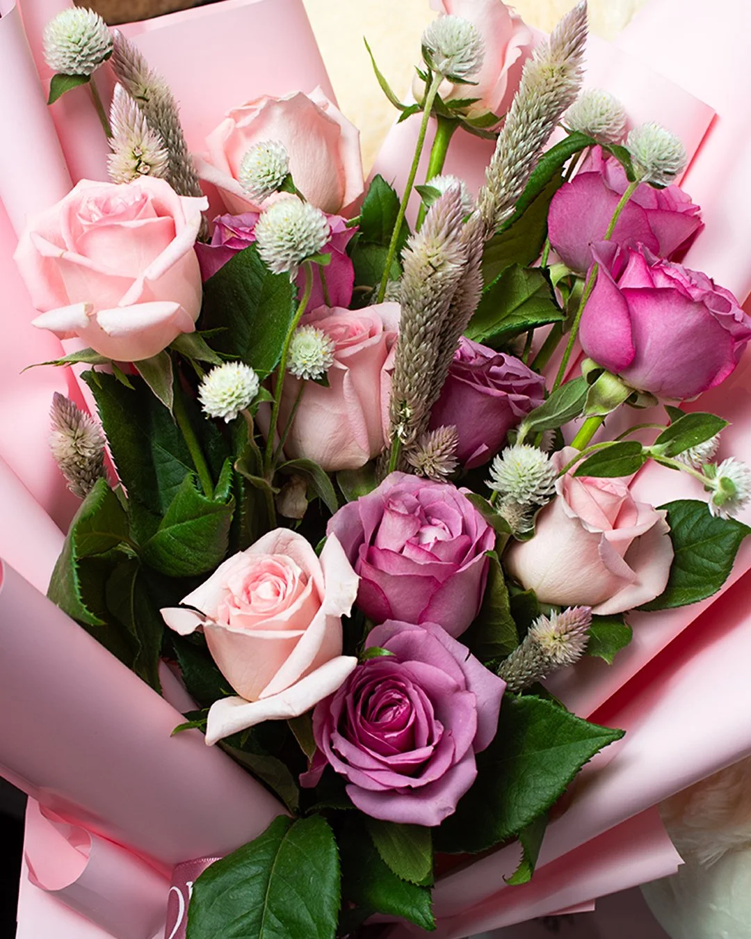 Close-up of a bouquet of pink and purple roses, white blooms, and green leaves wrapped in pink paper.