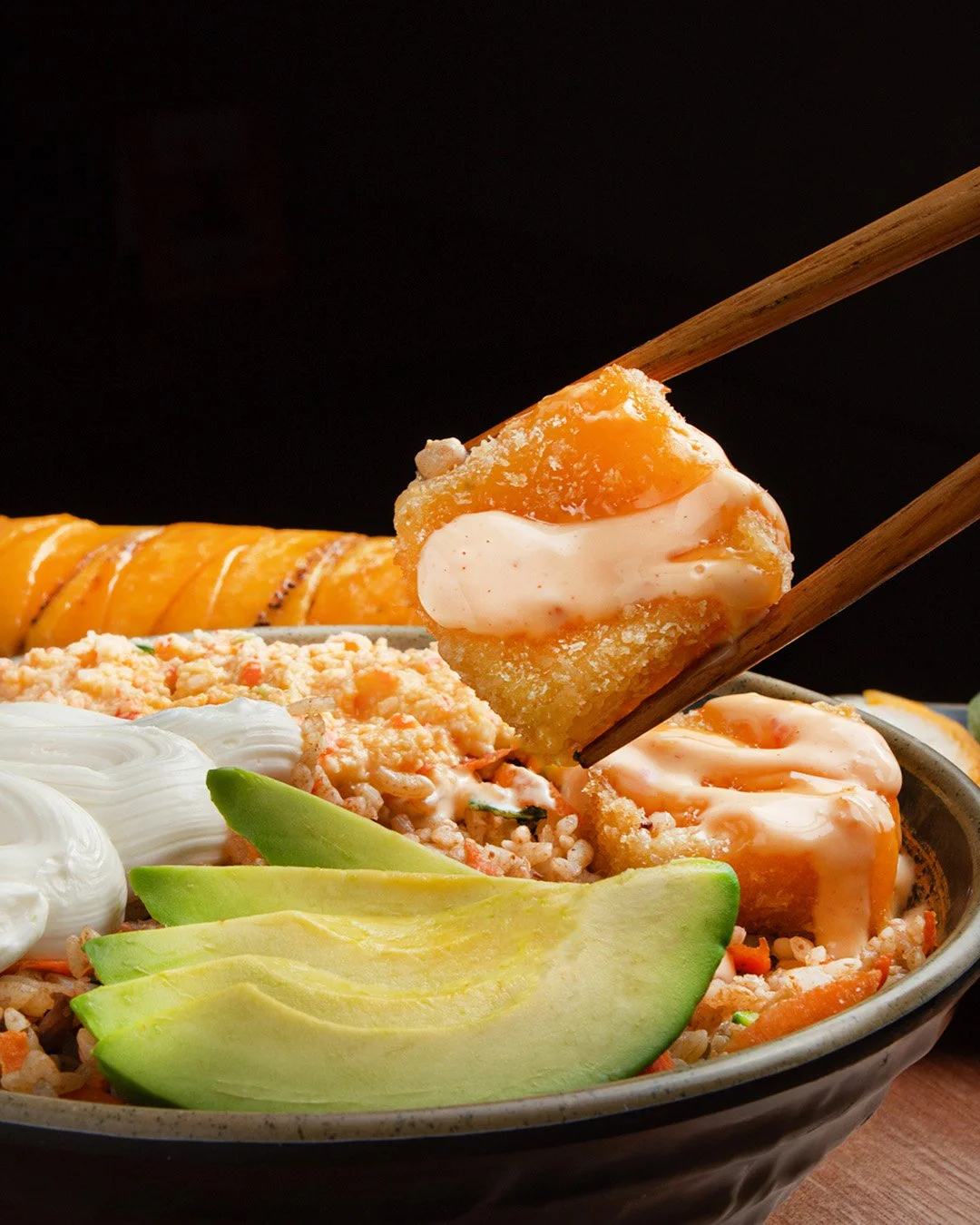 Close-up of a bowl of fried rice with avocado slices, sour cream, and some orange-colored sauce-covered fried food, possibly chicken or fish, being lifted with chopsticks against a black background.