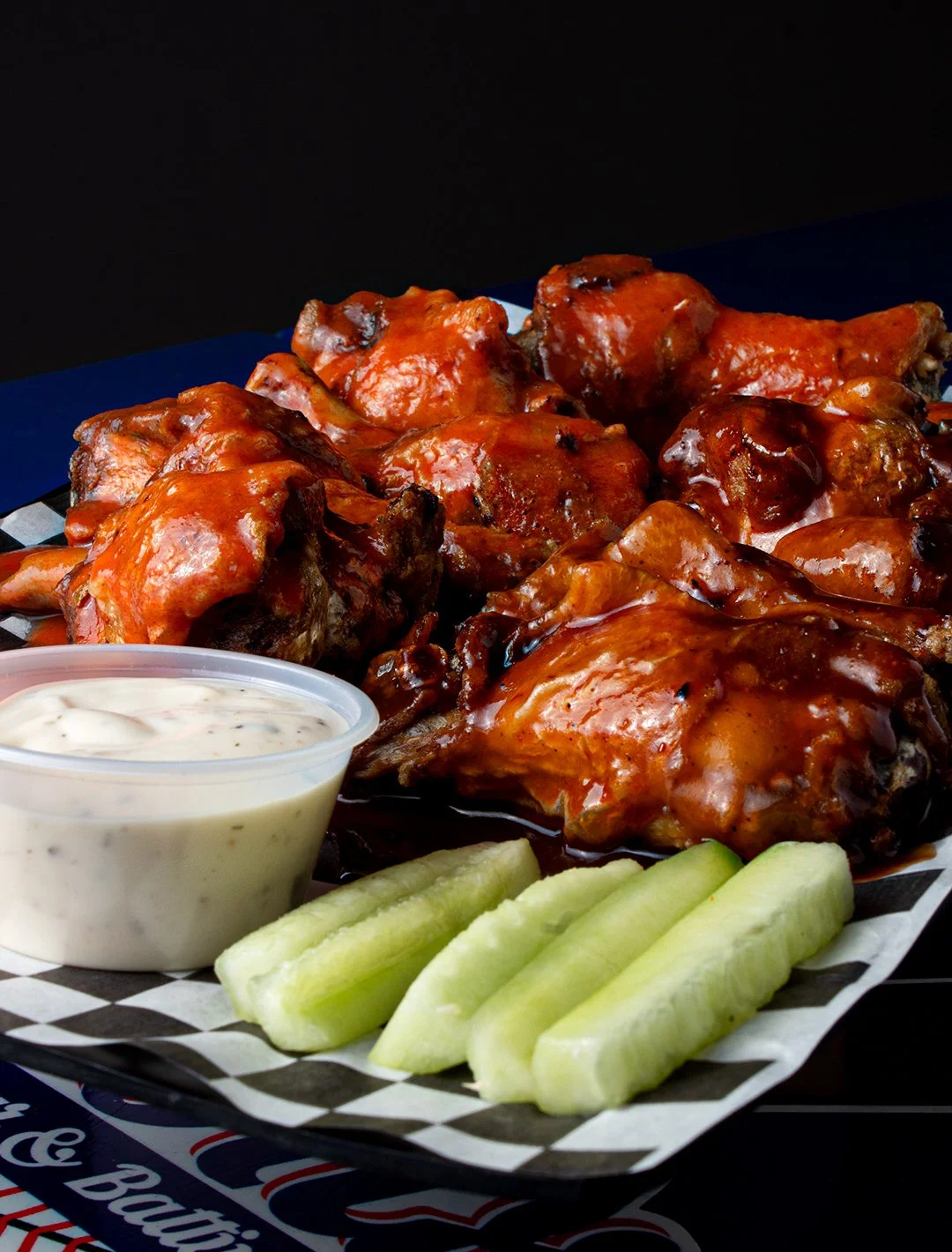 Plate of barbecue chicken drumsticks covered in barbecue sauce, side of cucumber slices, and a cup of ranch dressing, on black and white checkered paper liner.
