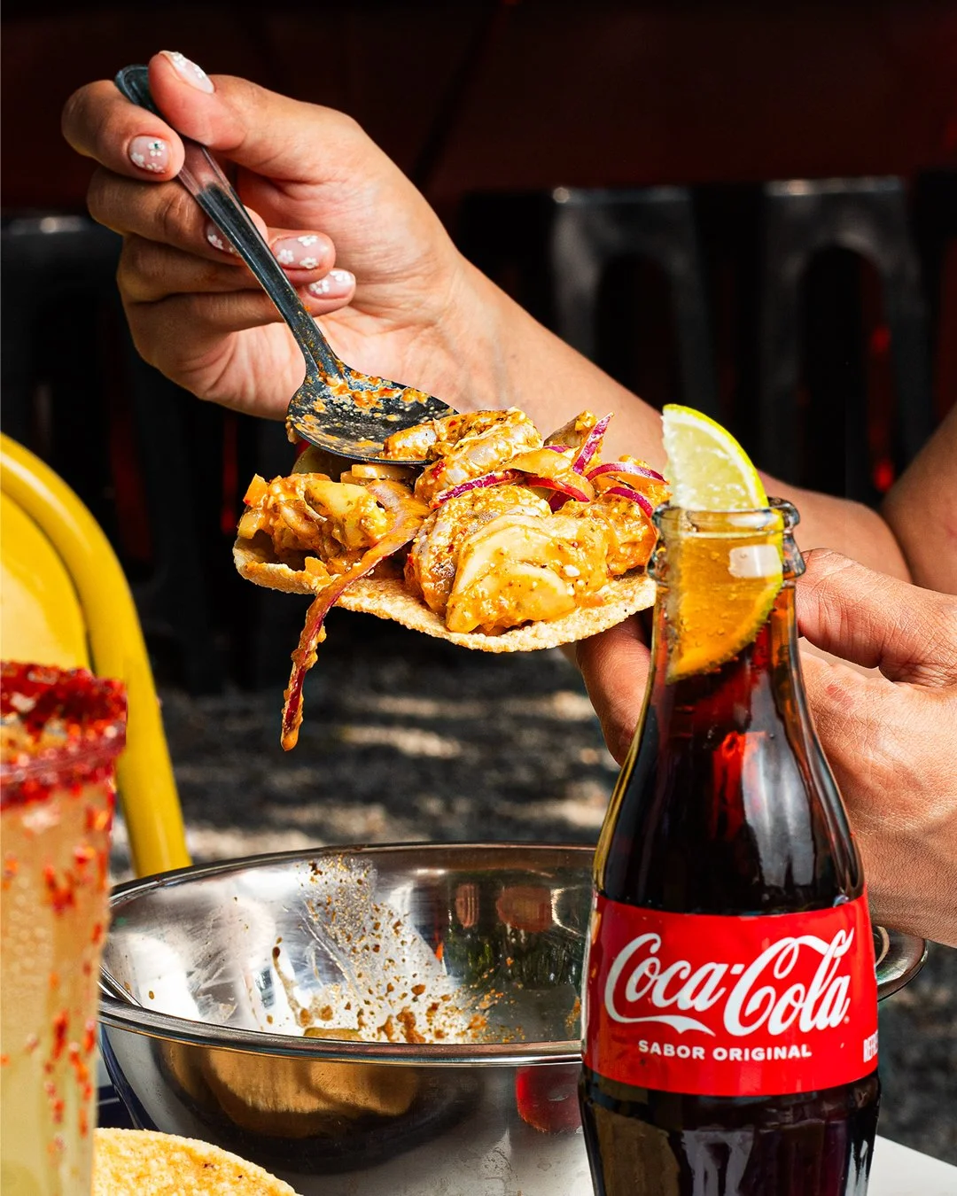 Person holding a spoon with a taco filled with seafood and garnished with onions and sauce, accompanied by a Coca-Cola soda with lemon on a table.