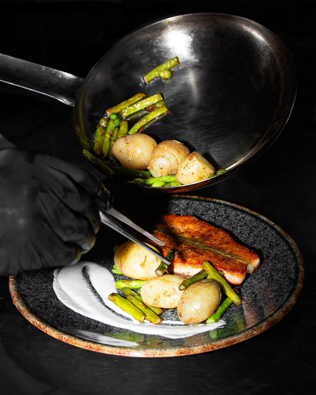 Chef plating cooked vegetables and a piece of fish on a black plate.