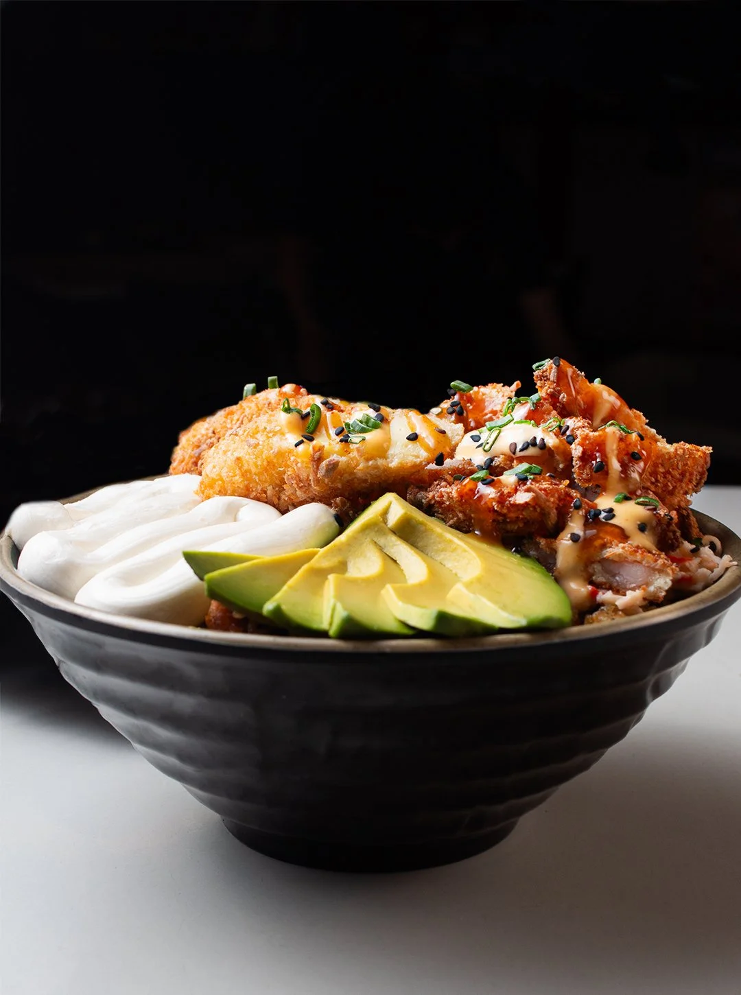 A black bowl filled with fried chicken topped with sauce and green onions, slices of avocado, and a dollop of sour cream or dressing.