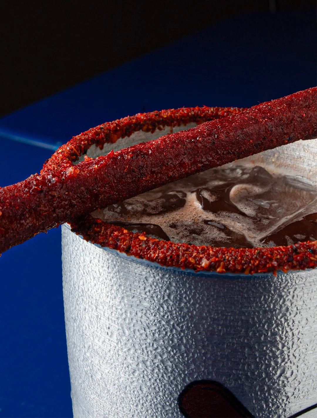 Frozen soda in a textured aluminum cup with a chili-rimmed straw resting on top, against a blue background.