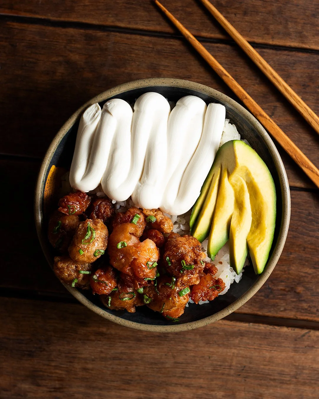 Bowl of sushi with sliced avocado, marinated chicken, and mayonnaise, on a wooden table with chopsticks.
