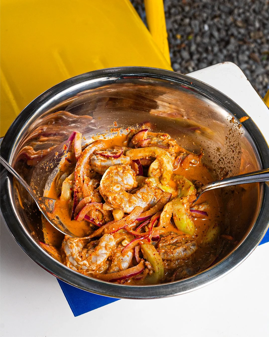 A metal bowl containing a mixed seafood and vegetable dish with a reddish sauce, placed on a white surface with a yellow background.