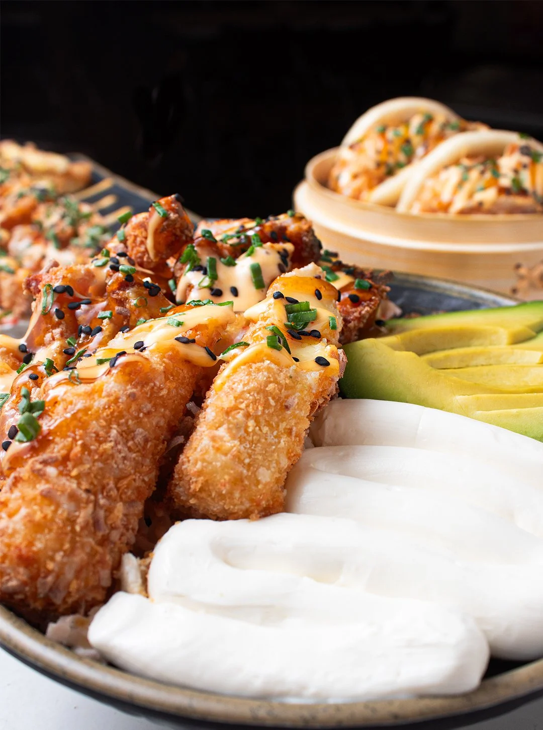 Close-up of a platter with fried sushi rolls topped with green onions, sesame seeds, and sauce, alongside slices of avocado and white fish, with a bamboo steamer containing more sushi rolls in the background.