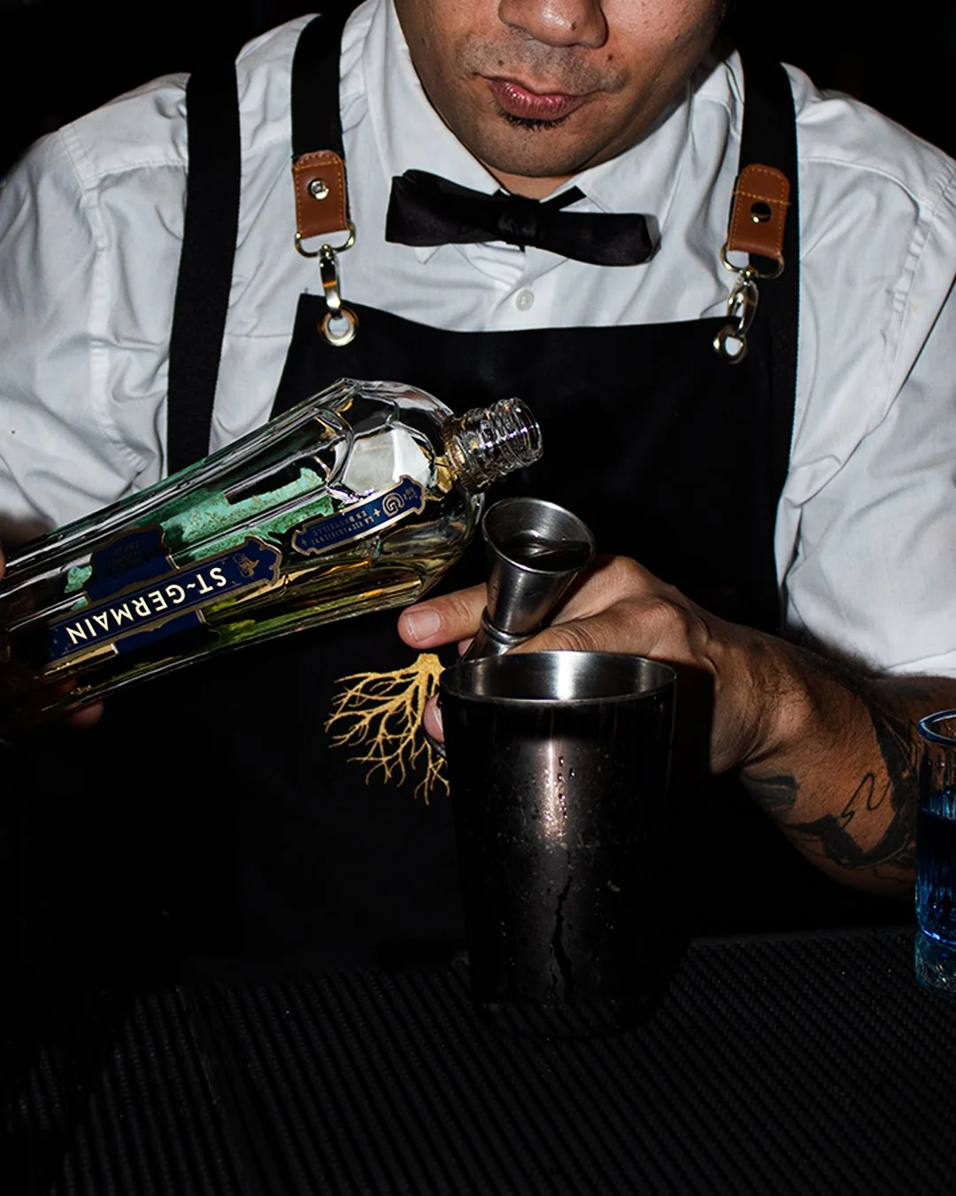 Bartender pouring Grey Goose vodka into a metal cocktail shaker, wearing a white shirt, black bow tie, and black apron.