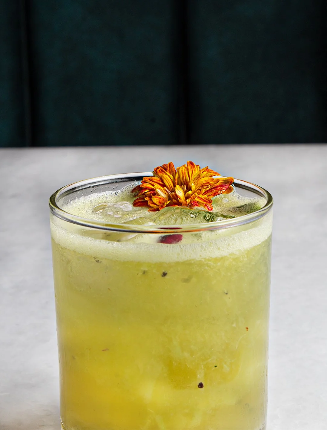 Yellow cocktail in a glass garnished with an orange flower, on a white surface with a dark background.